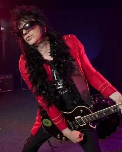 A musician dressed in a red jacket, black shirt, and sunglasses, holding a black electric guitar on stage with dark lighting.