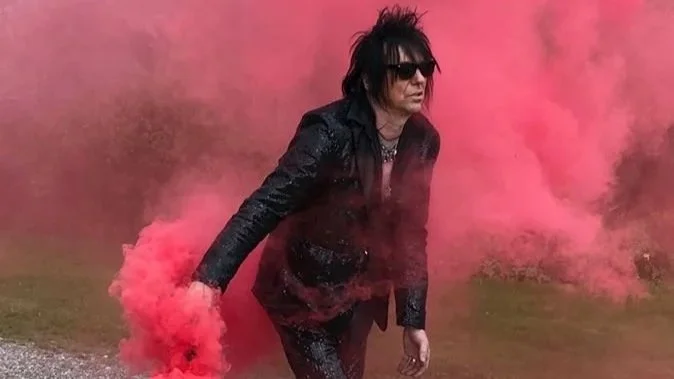 A man dressed in black with sunglasses and spiky dark hair walking through pink smoke.