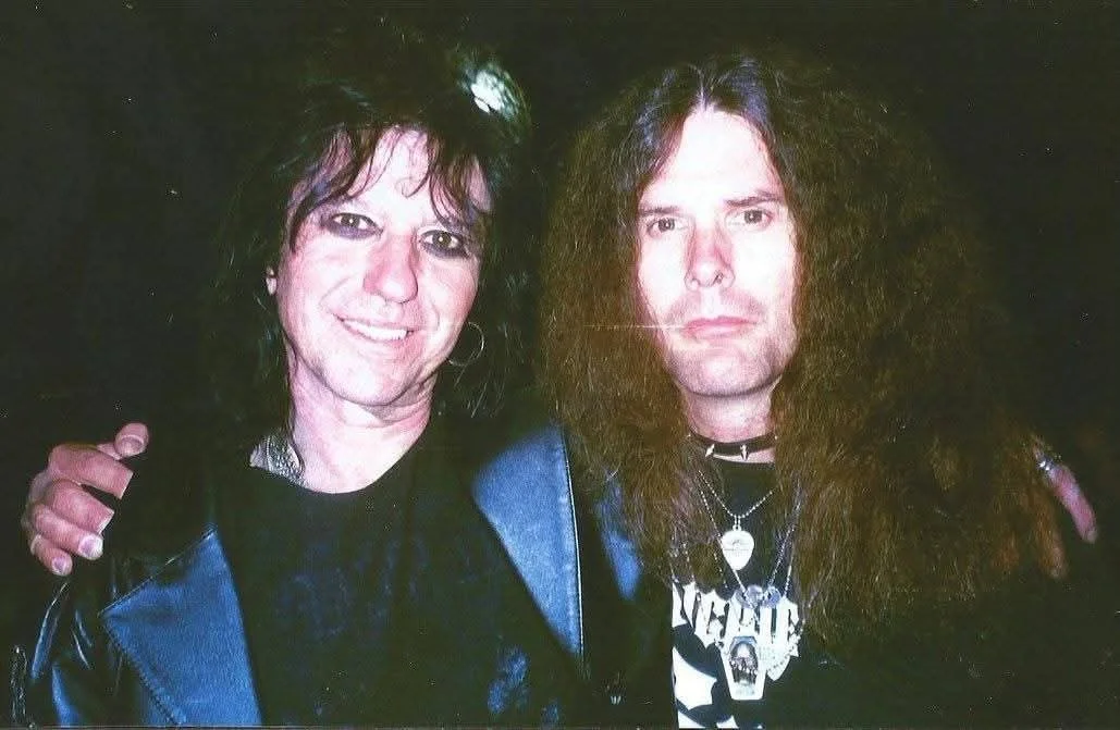 Two men with long dark hair, wearing black clothing, standing close together with arms around each other in a dark setting.