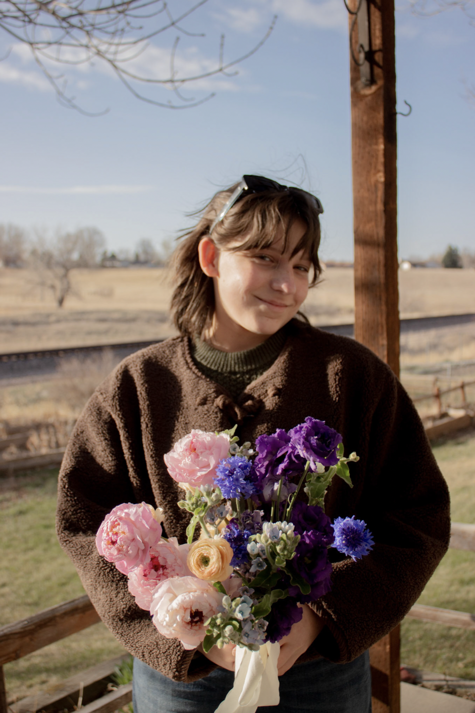 A young woman with shoulder-length brown hair holding a bouquet of pink, purple, and blue flowers, standing outdoors on a sunny day.