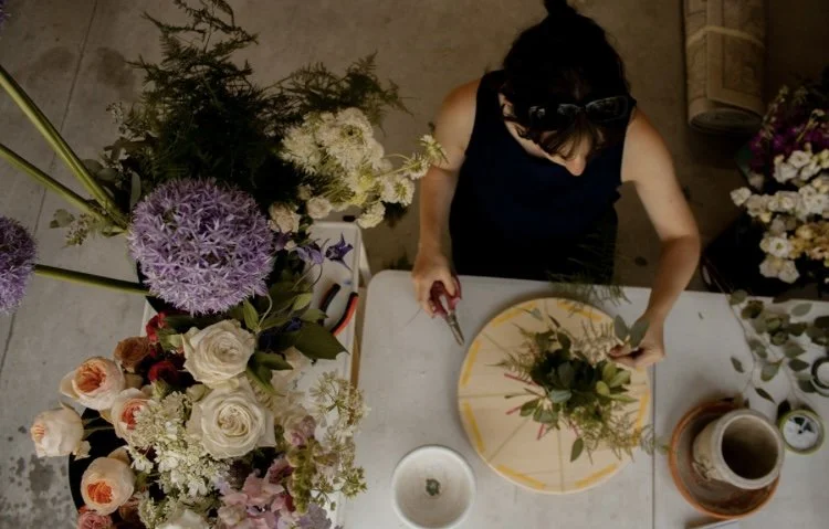 A woman in a dark sleeveless top arranging flowers on a round floral design on a table, surrounded by various bouquets of flowers.