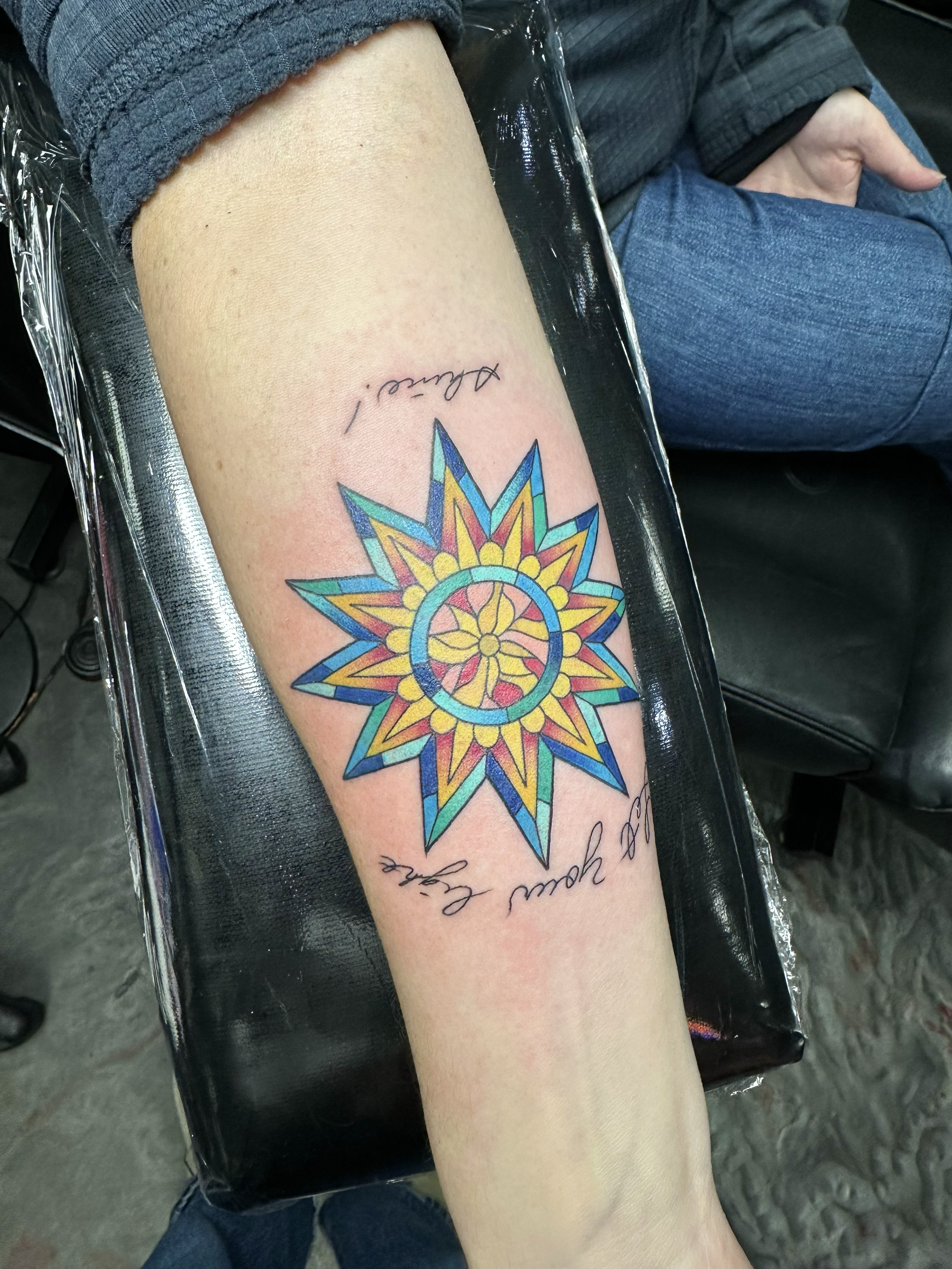 Colorful tattoo of a starburst design with a peace symbol in the center on a person's forearm, with script text above and below the tattoo.