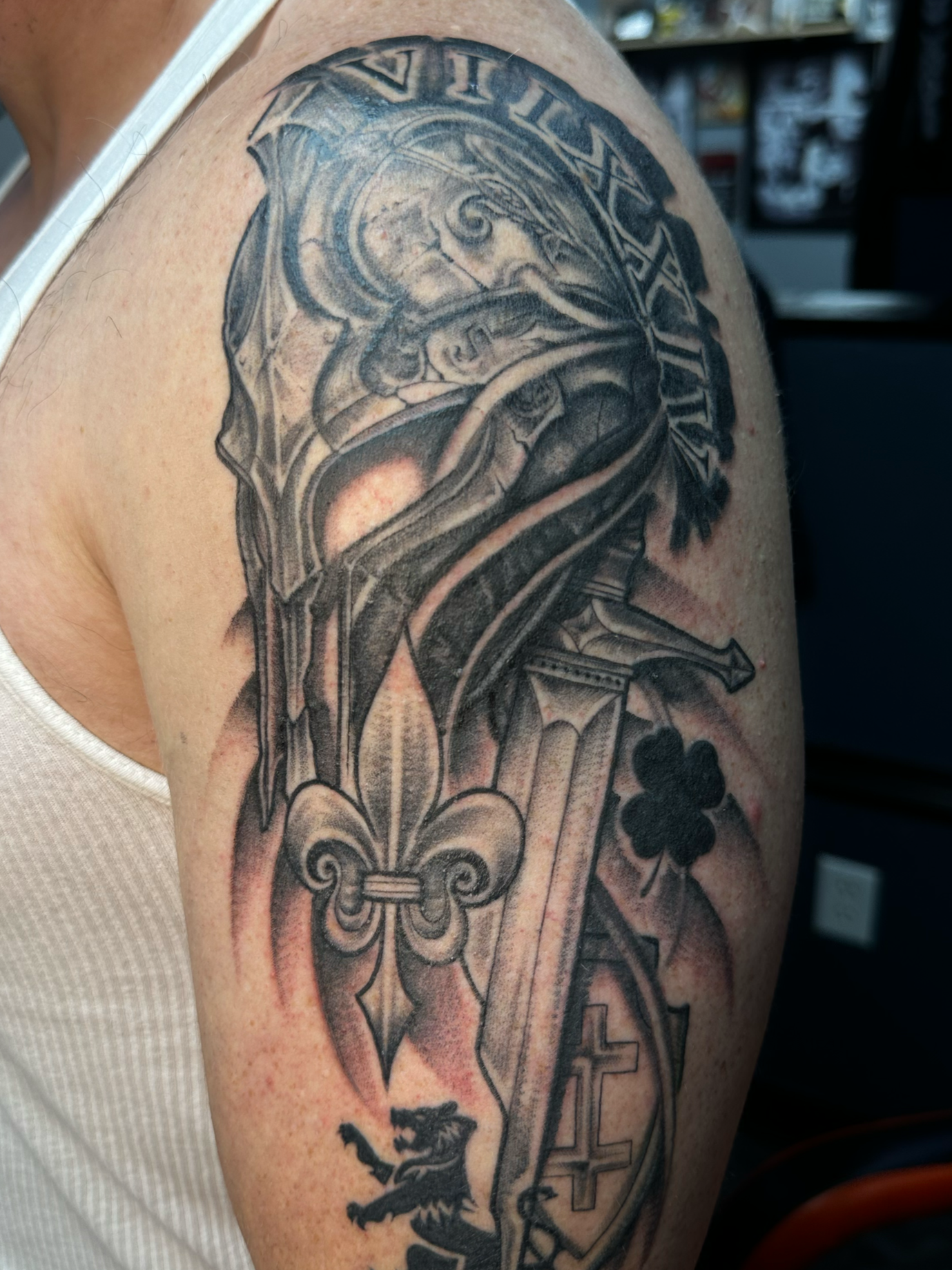 Black and gray tattoo on upper arm features a dragon with a shield, a sword, a three-leaf clover, a lion, and the words 'VIVAT MILITIS'.