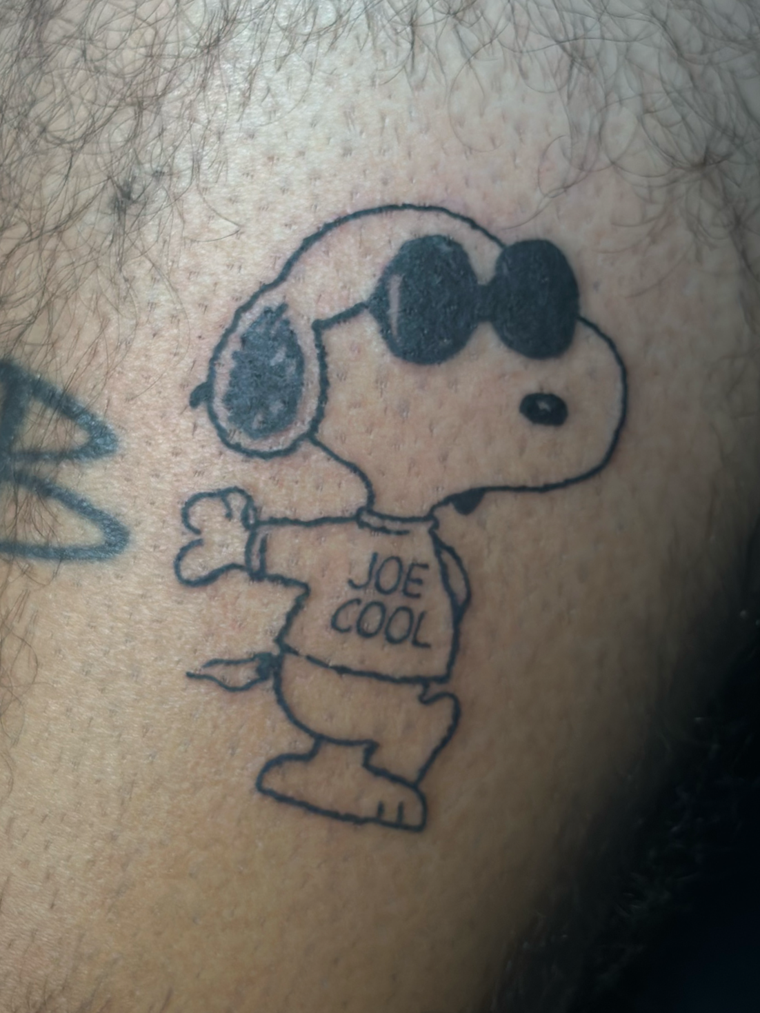 Close-up of a tattoo of Snoopy wearing sunglasses and a t-shirt that says 'JOE COOL' on an arm with hair.