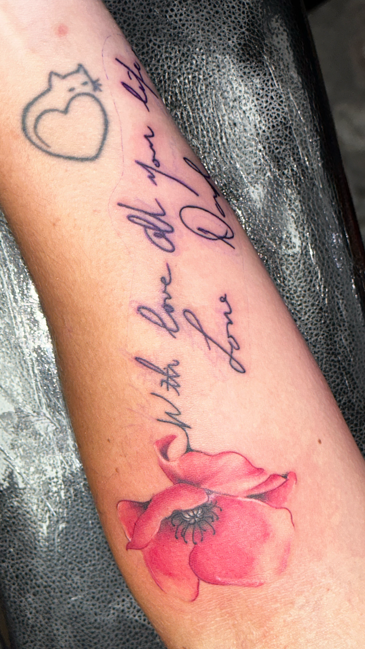 Tattoo of a red poppy flower with black details and a black outline on a person's forearm, accompanied by cursive writing that says 'You will love again and again' and a small heart with a star.
