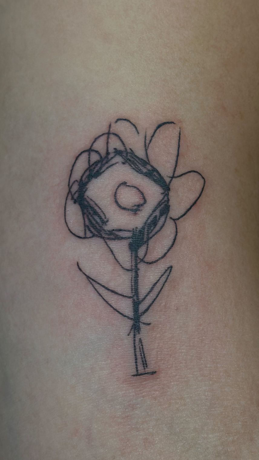 A black ink tattoo of a simple, abstract flower on skin. The drawing is rough with irregular, sketchy lines.