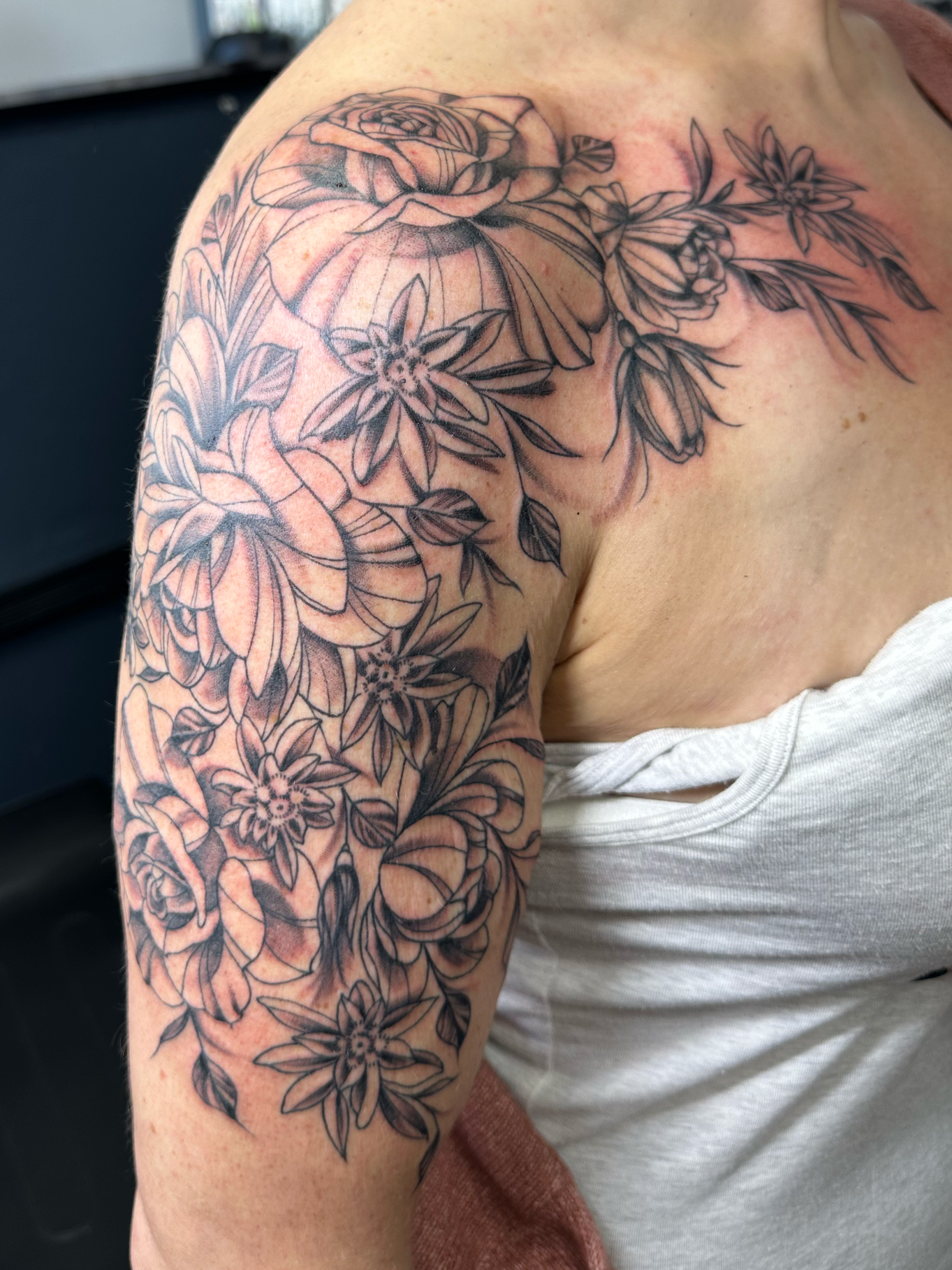 Black outline floral tattoo on person's shoulder and upper arm, featuring various flowers and leaves.