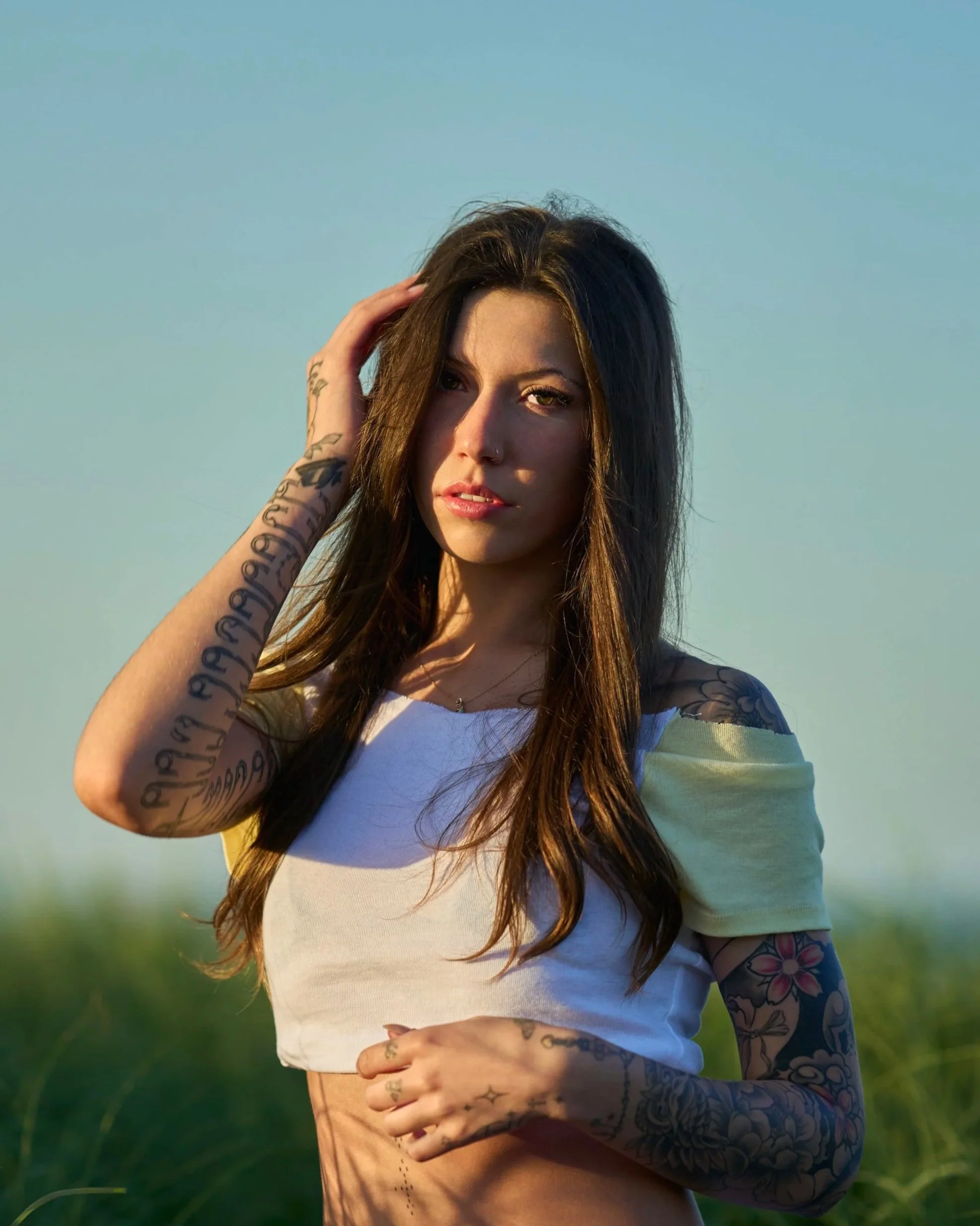 A young woman with long dark hair and visible tattoos, standing outdoors during sunset with a blue sky, touching her hair with one hand and resting the other on her waist.