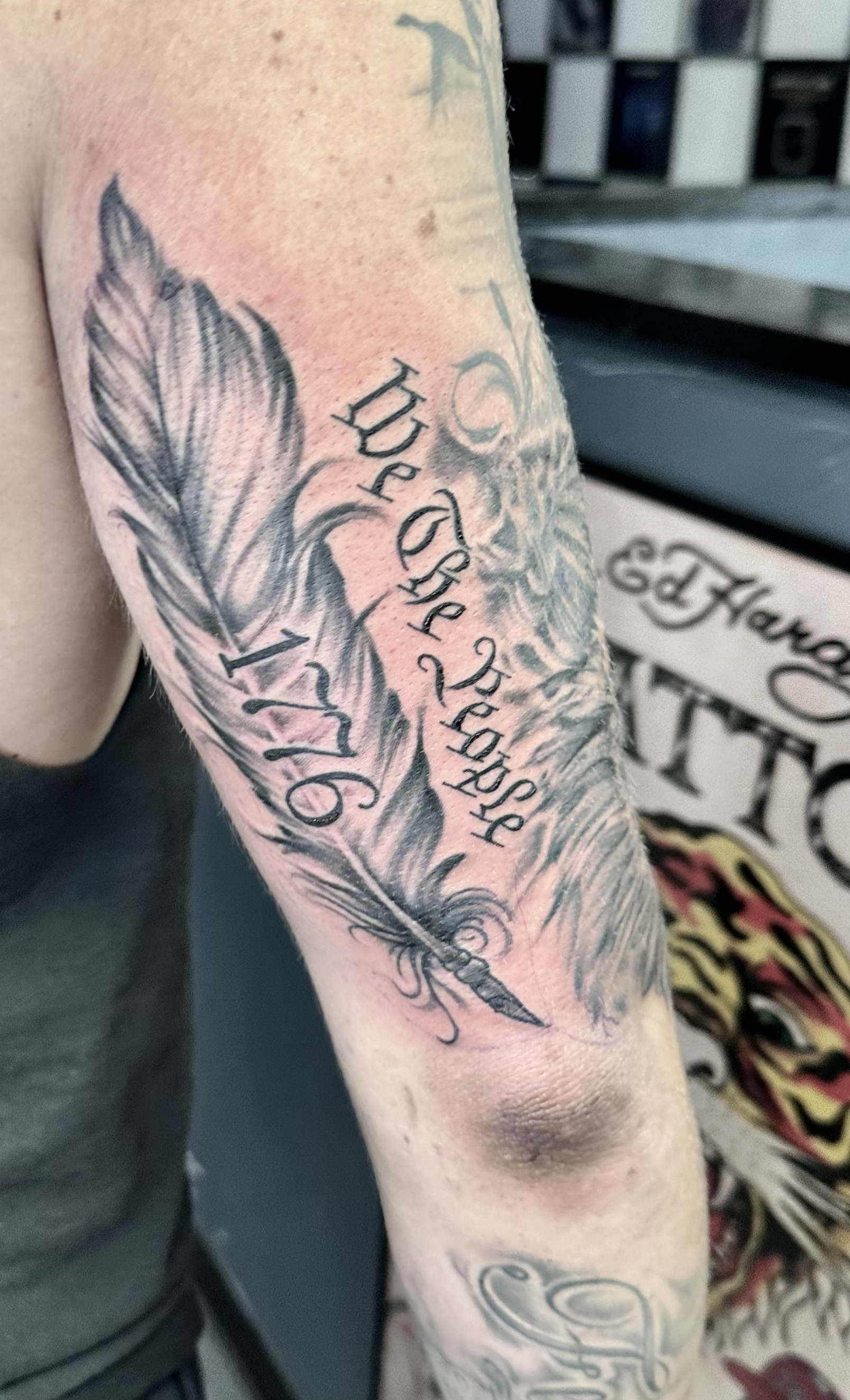 Tattoo of a feather with cursive text that reads "Hispanic" and "1976" on a person's arm.