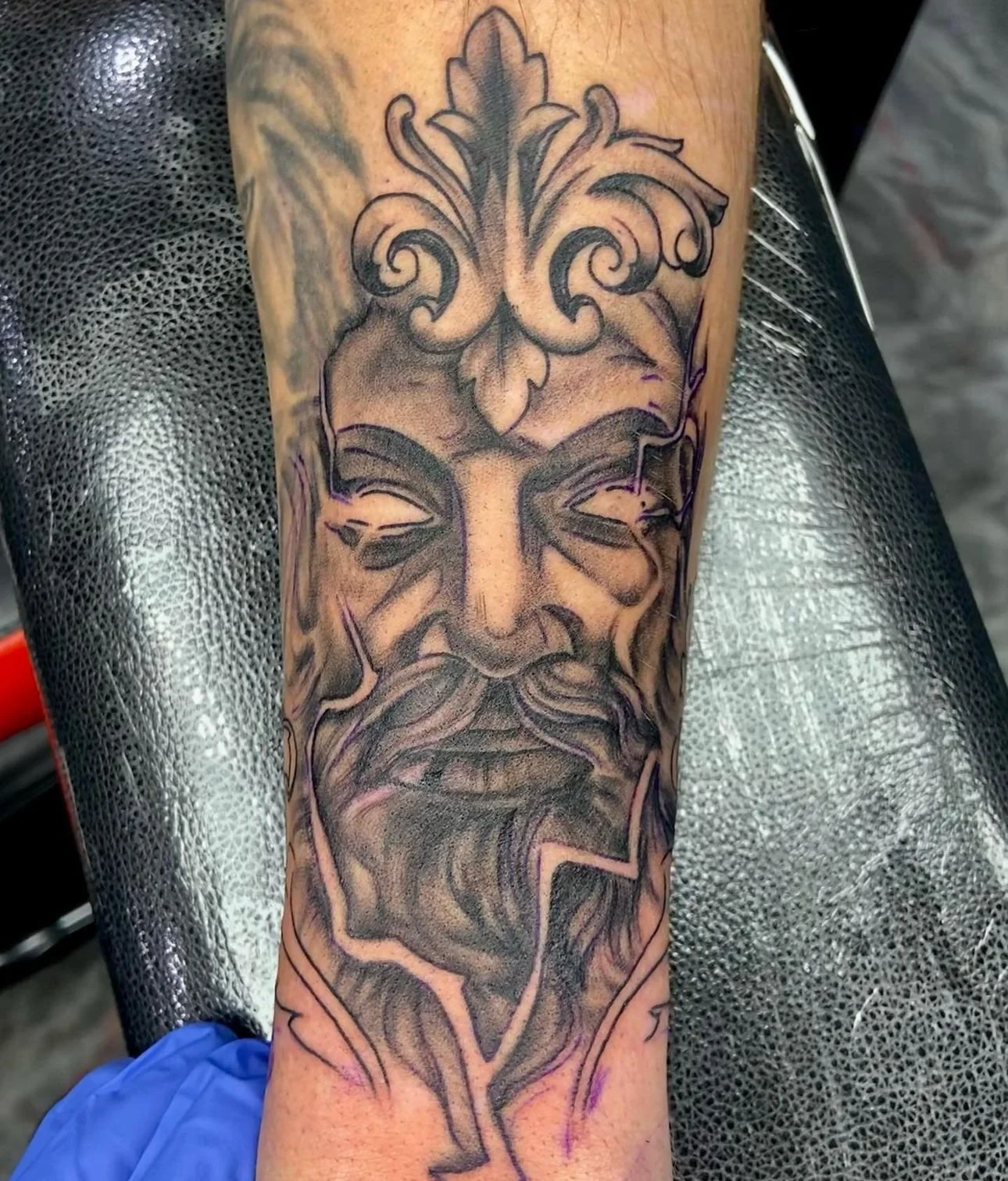 A tattoo of a face with closed eyes and a flowing beard, framed by decorative floral patterns and ornate details, in black and gray ink.