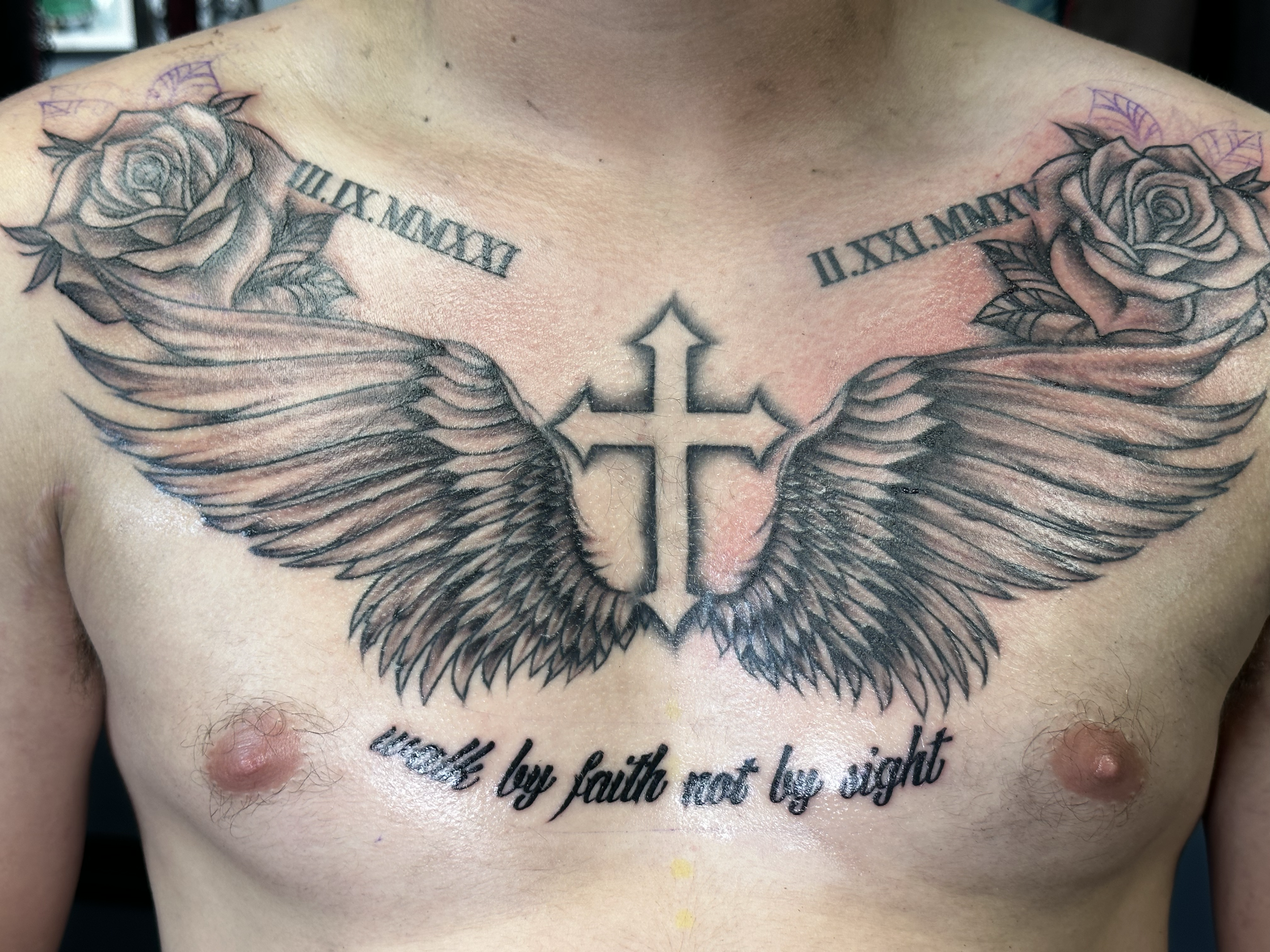Tattoo of a cross with angel wings, roses, and a phrase on a person's chest.