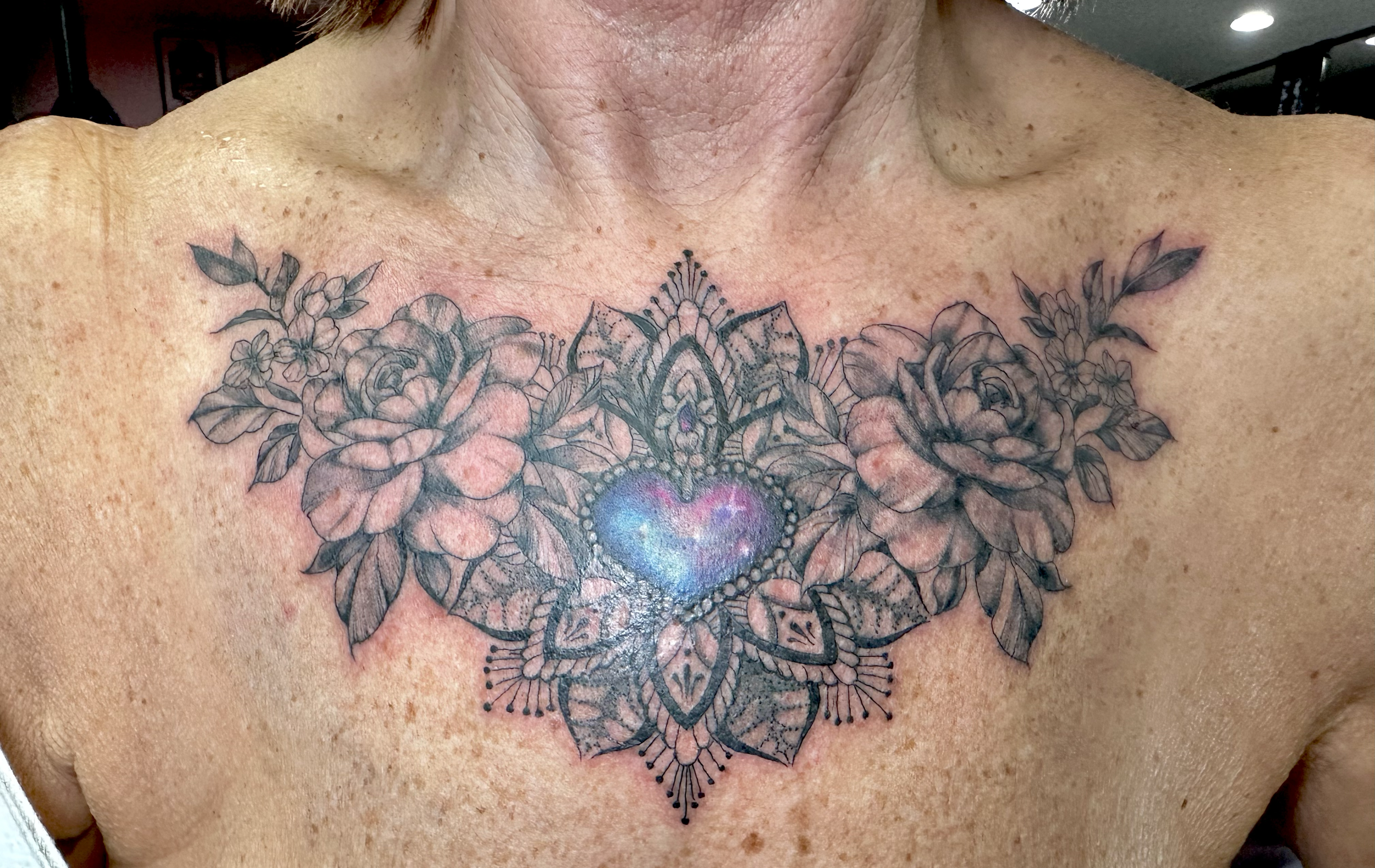 Tattoo of a heart surrounded by detailed floral and geometric designs on a person's chest.