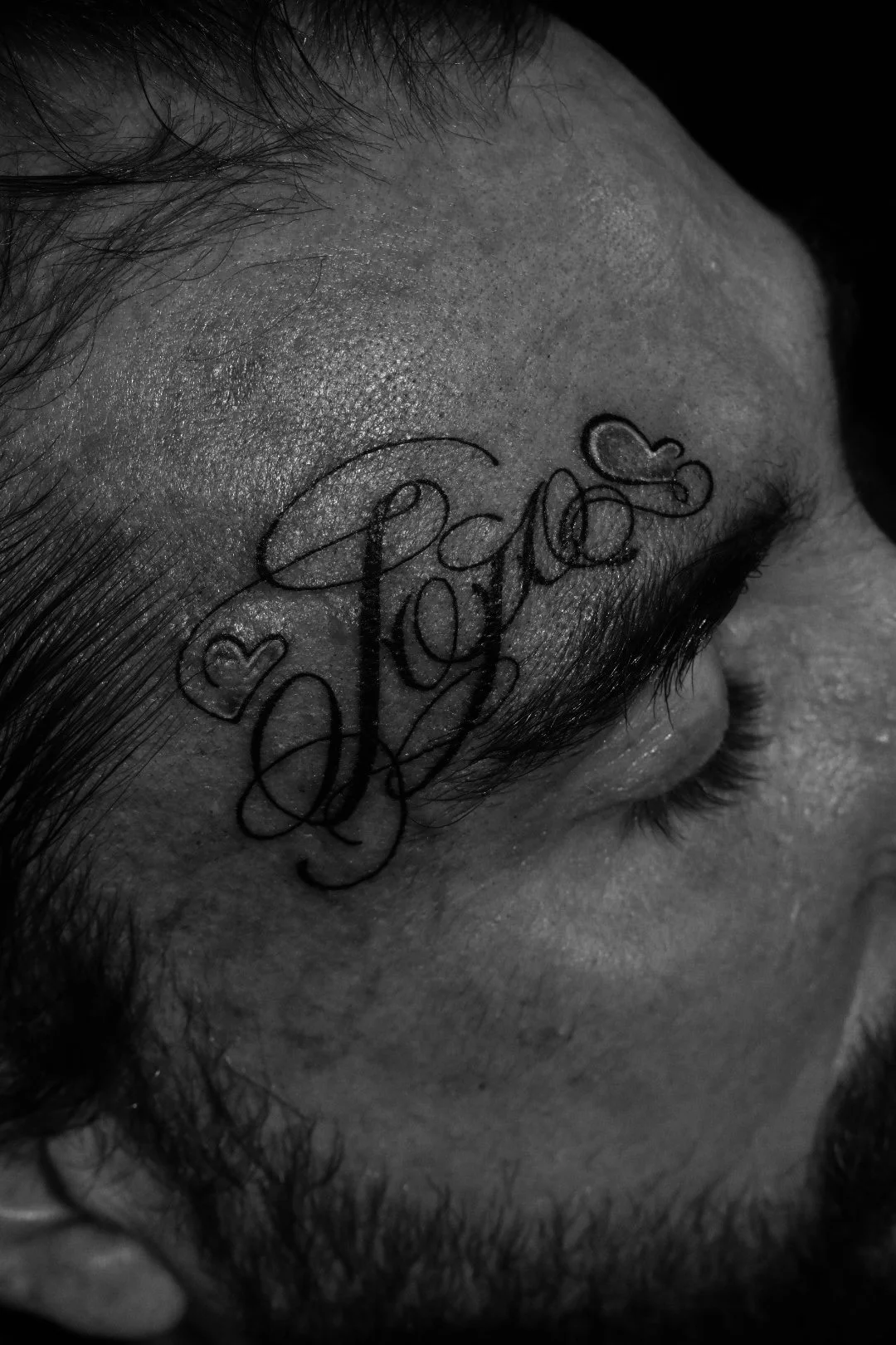 Close-up of a person's forehead and eye with a tattoo that reads 'Love' in cursive, surrounded by small hearts, in black and white.
