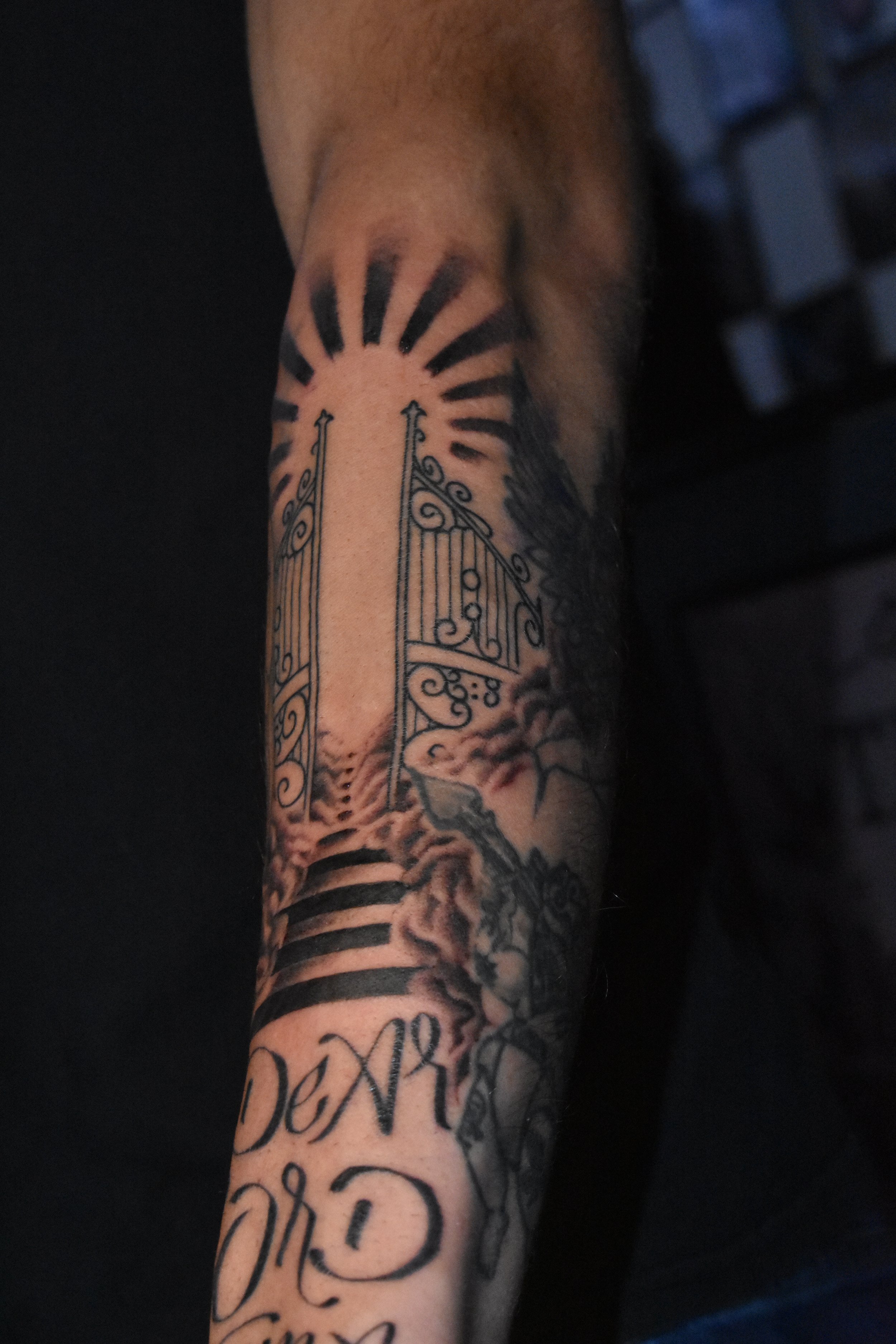 Tattooed arm featuring a gate opening to a landscape with clouds and rays of light, with decorative elements and script.
