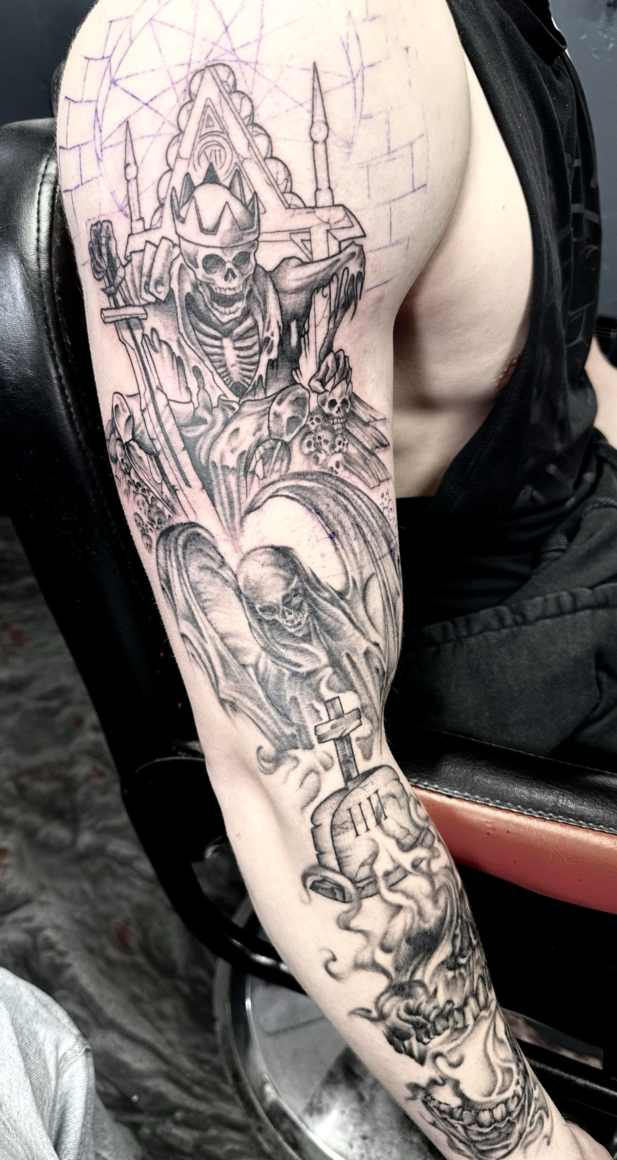 Black and gray tattoo sleeve featuring a skull-faced skeleton king with a crown, surrounded by skulls and weapons, a figure with a sword, a tombstone with a cross, and a demonic creature with horns.