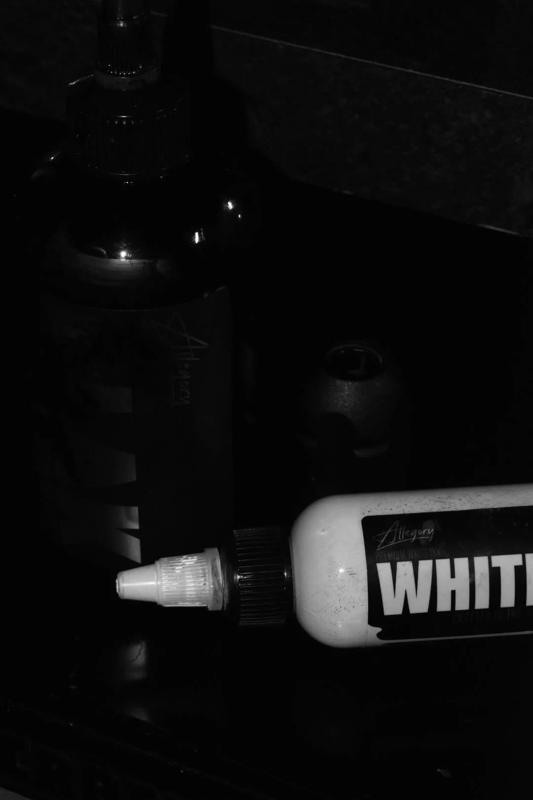 A bottle of white liquid allergy medication lying on its side with a dispensing nozzle, a partially visible soda can, a dark spray bottle, and an unknown object in a dark environment.