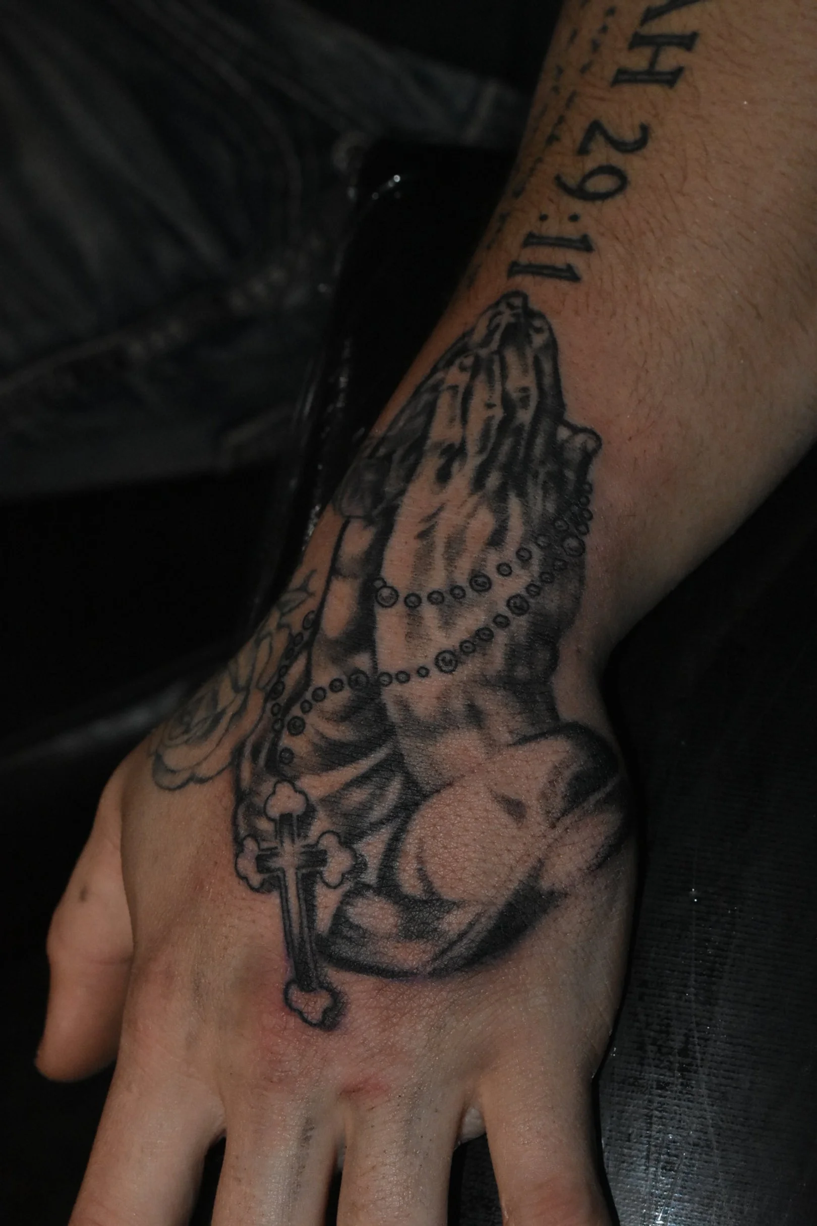 Tattoo of praying hands holding a rosary with a cross, programmatically minified and part of a colorful arm tattoo.