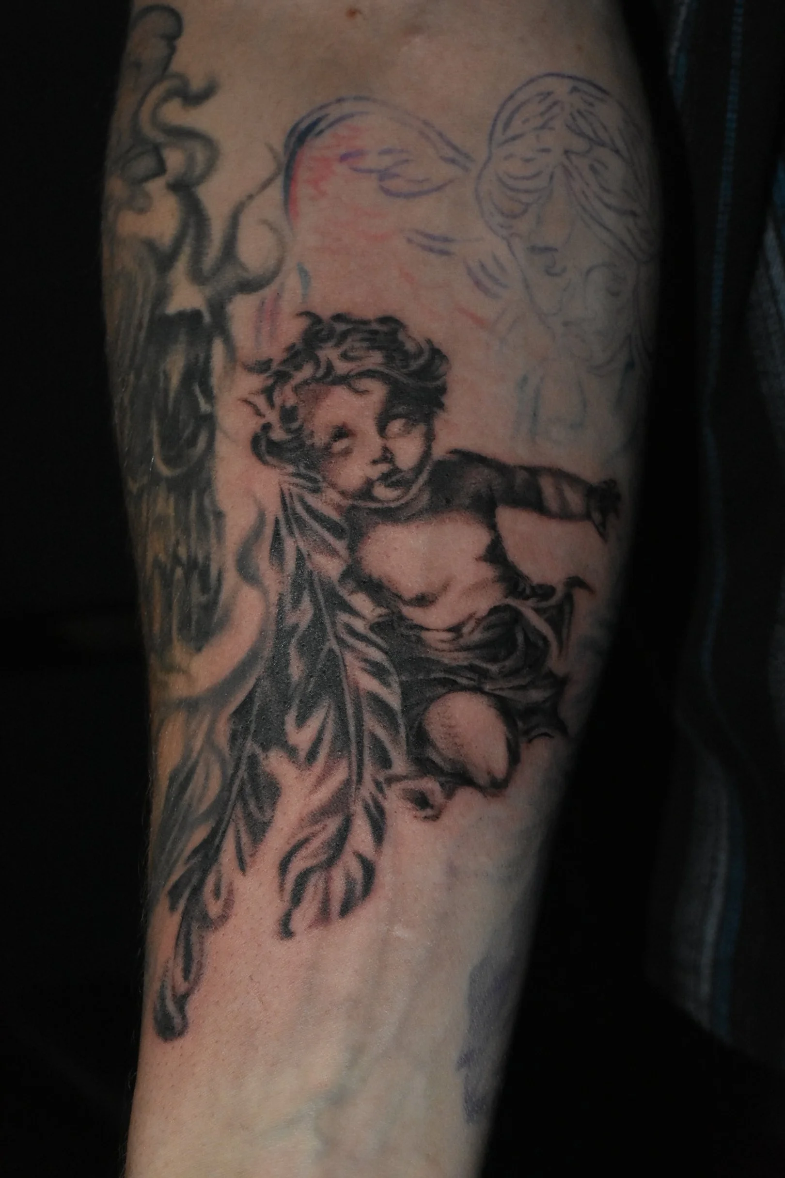 Black and gray tattoo of a young boy with curly hair, partially nude, holding a large feather, with sketches of angelic faces in the background.