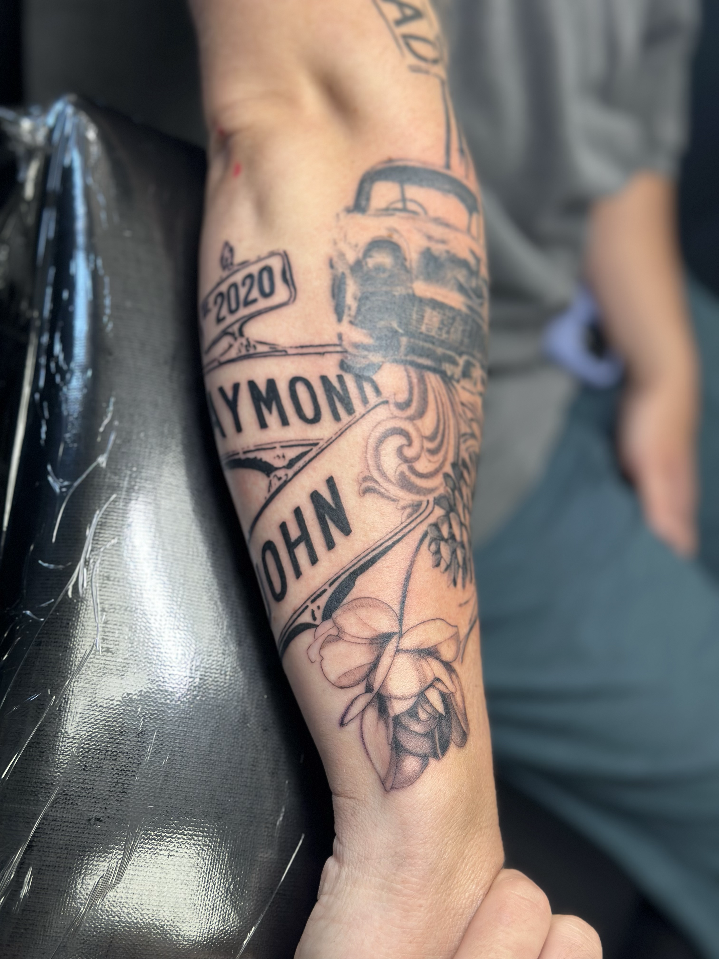 Tattoo sleeve on person's arm featuring a train, banners with the year 2020, the name Yvonne, the name John, and a butterfly.
