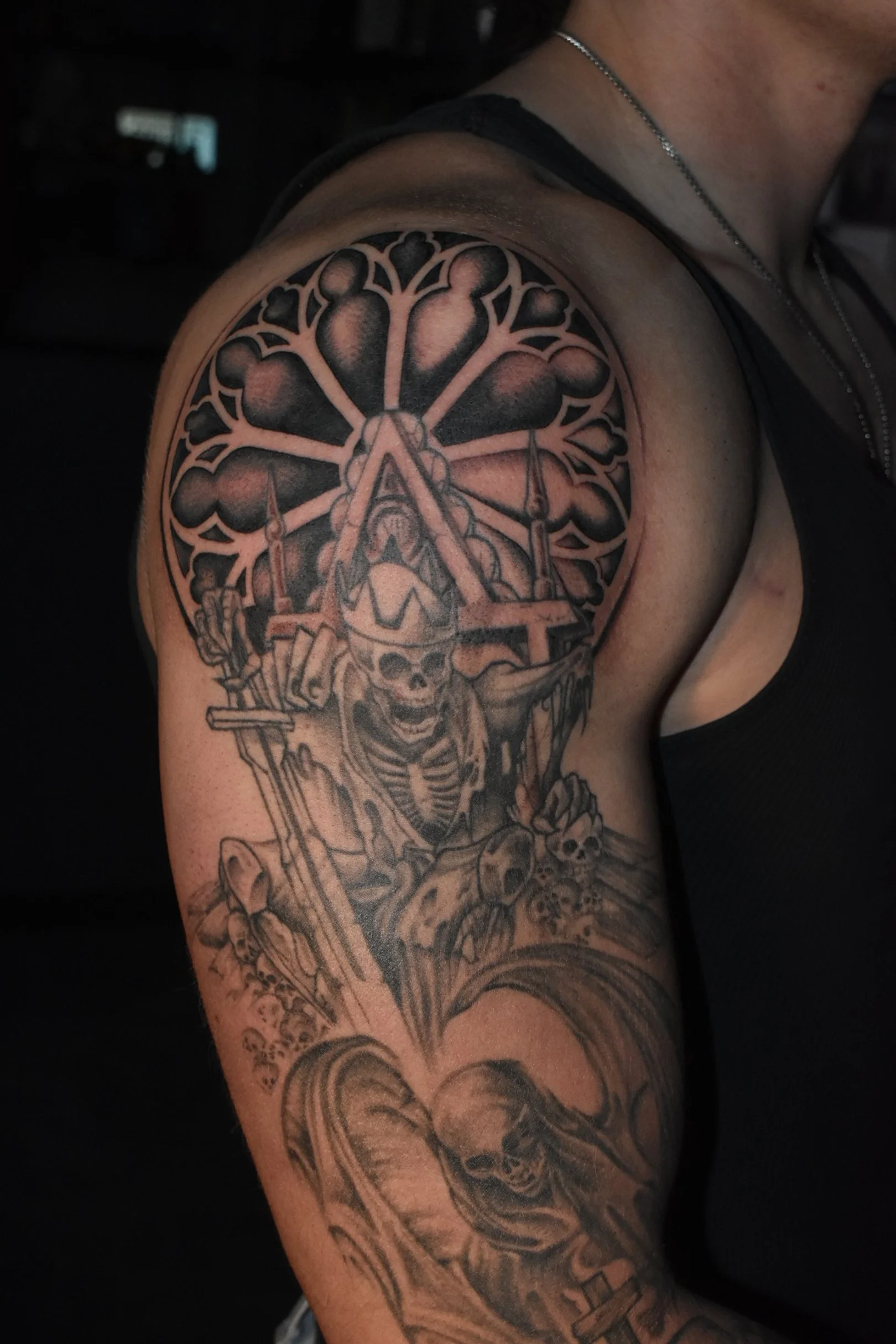 Black and grey tattoo on a person's upper arm featuring a skeleton wearing a helmet with goggles, surrounded by skulls, bones, and a large tree with many branches and heart-shaped leaves.