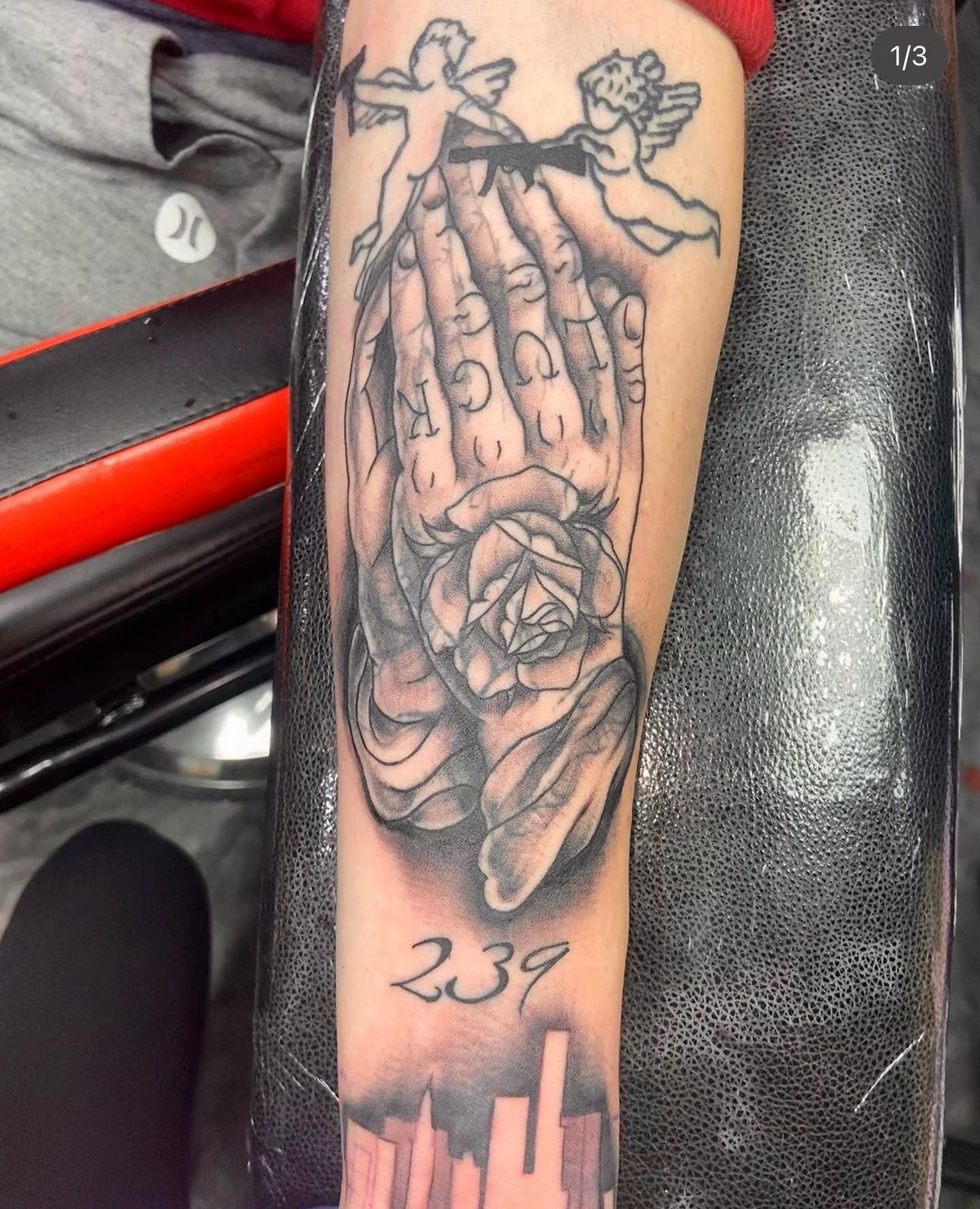 Tattoo on a person's forearm depicting a hand holding a rose with two cherubs flying above and the number 239 below.