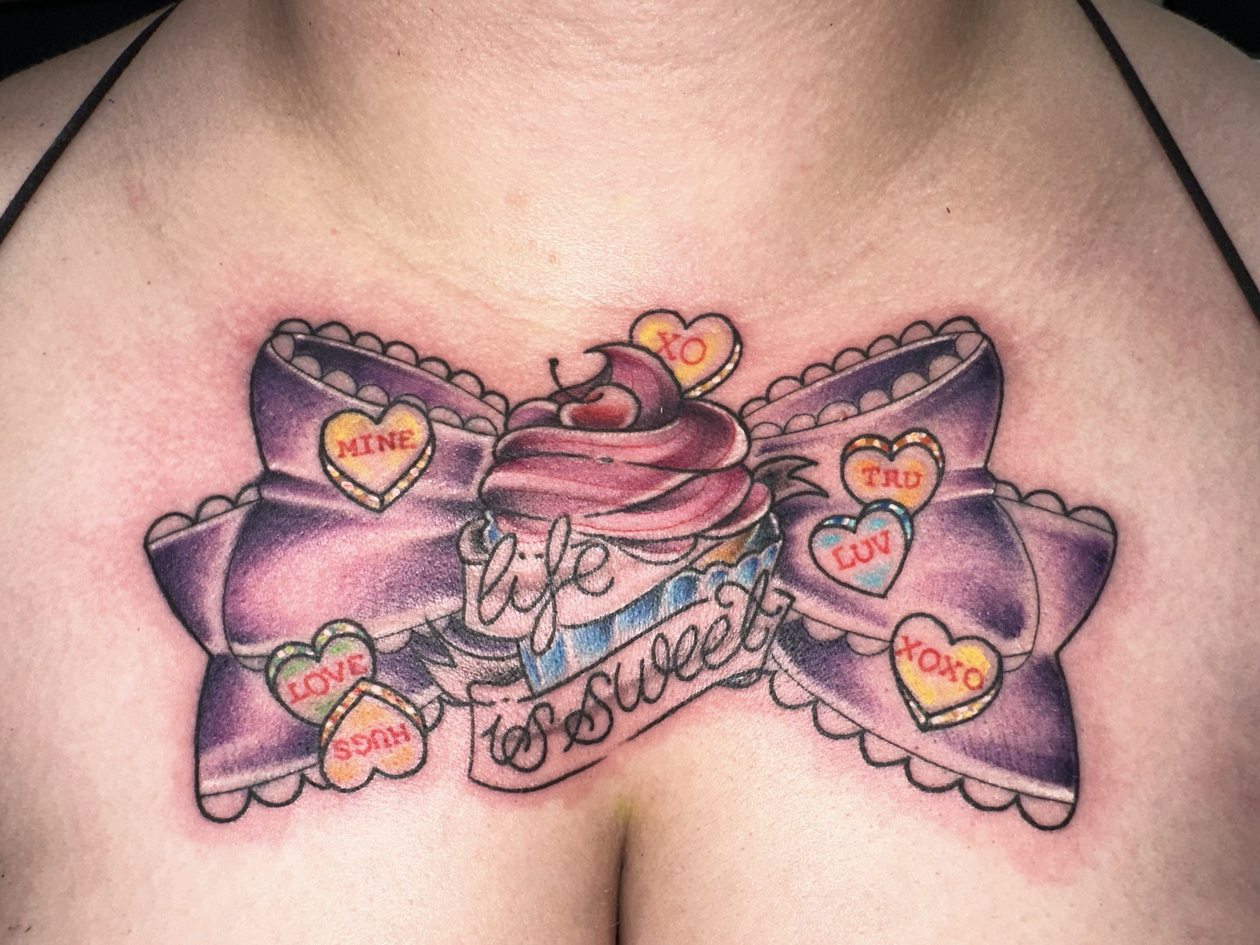 Tattoo of a cupcake with purple icing and red cherry on top, surrounded by a purple bow with hearts containing words like 'mine,' 'love,' 'trust,' 'xoxo,' and the phrase 'life is sweet.'