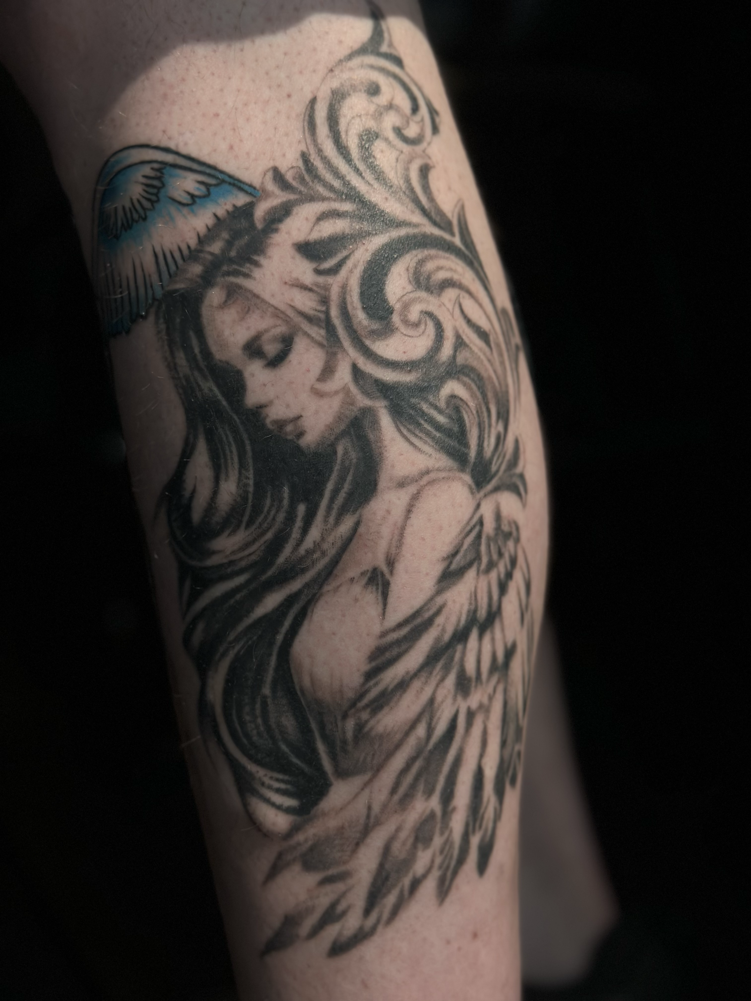 Tattoo of a woman with long flowing hair, with angel wings, and a blue and black feathered wing, on a person's arm.
