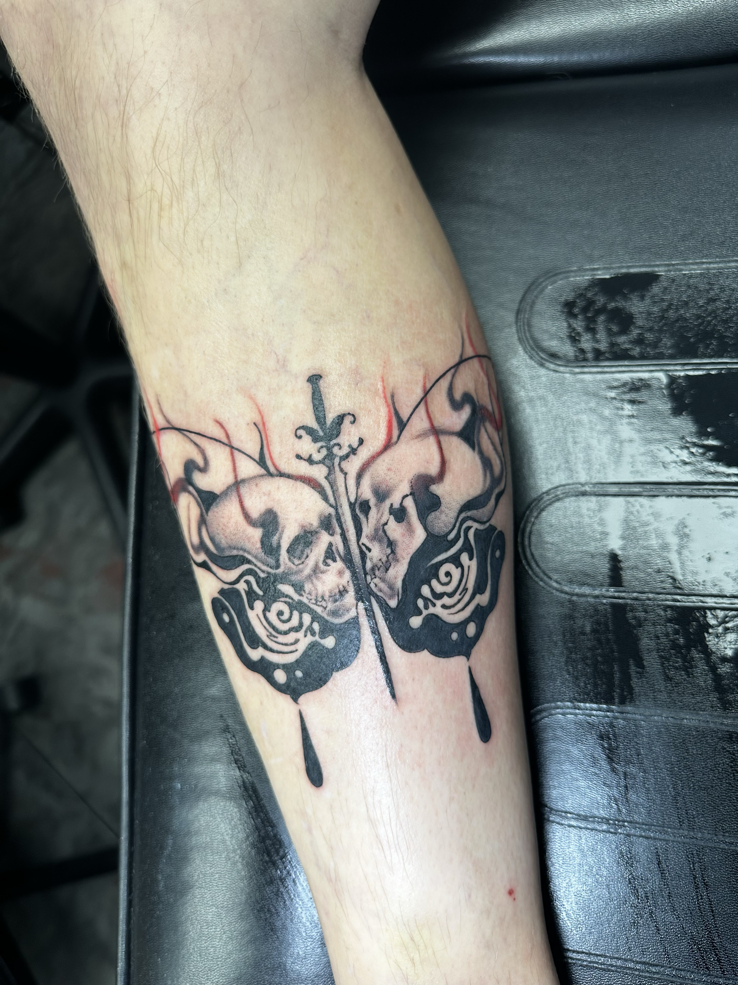 A tattoo of a butterfly with skulls on the sides and a sword through the middle, with black ink and red accents, on a person's leg.