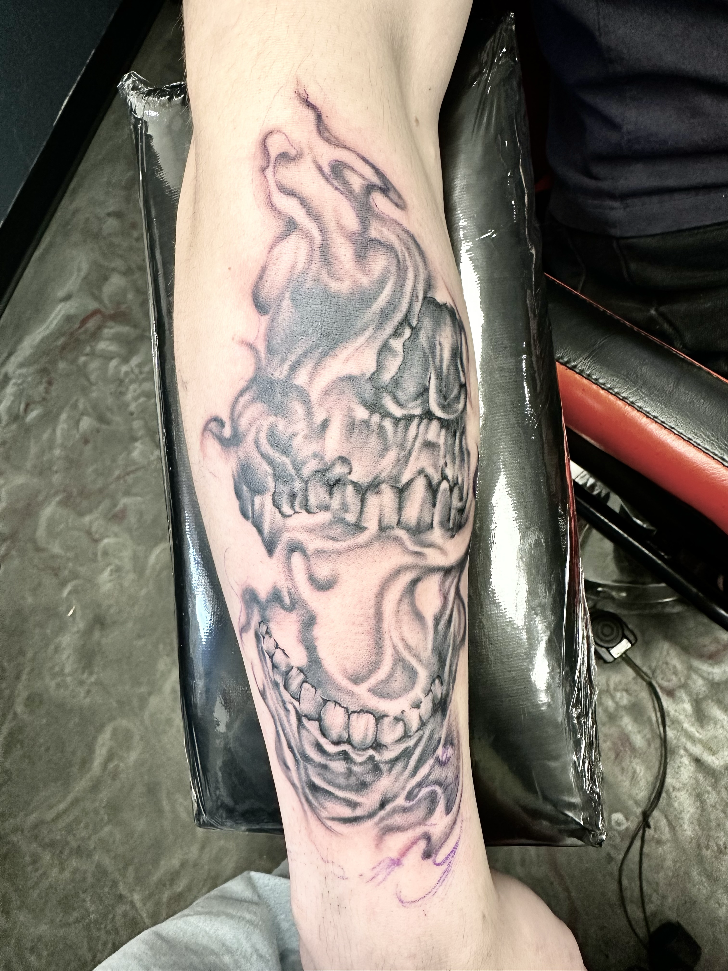 A black and gray tattoo of a skull with smoke swirling around it on a person's forearm.