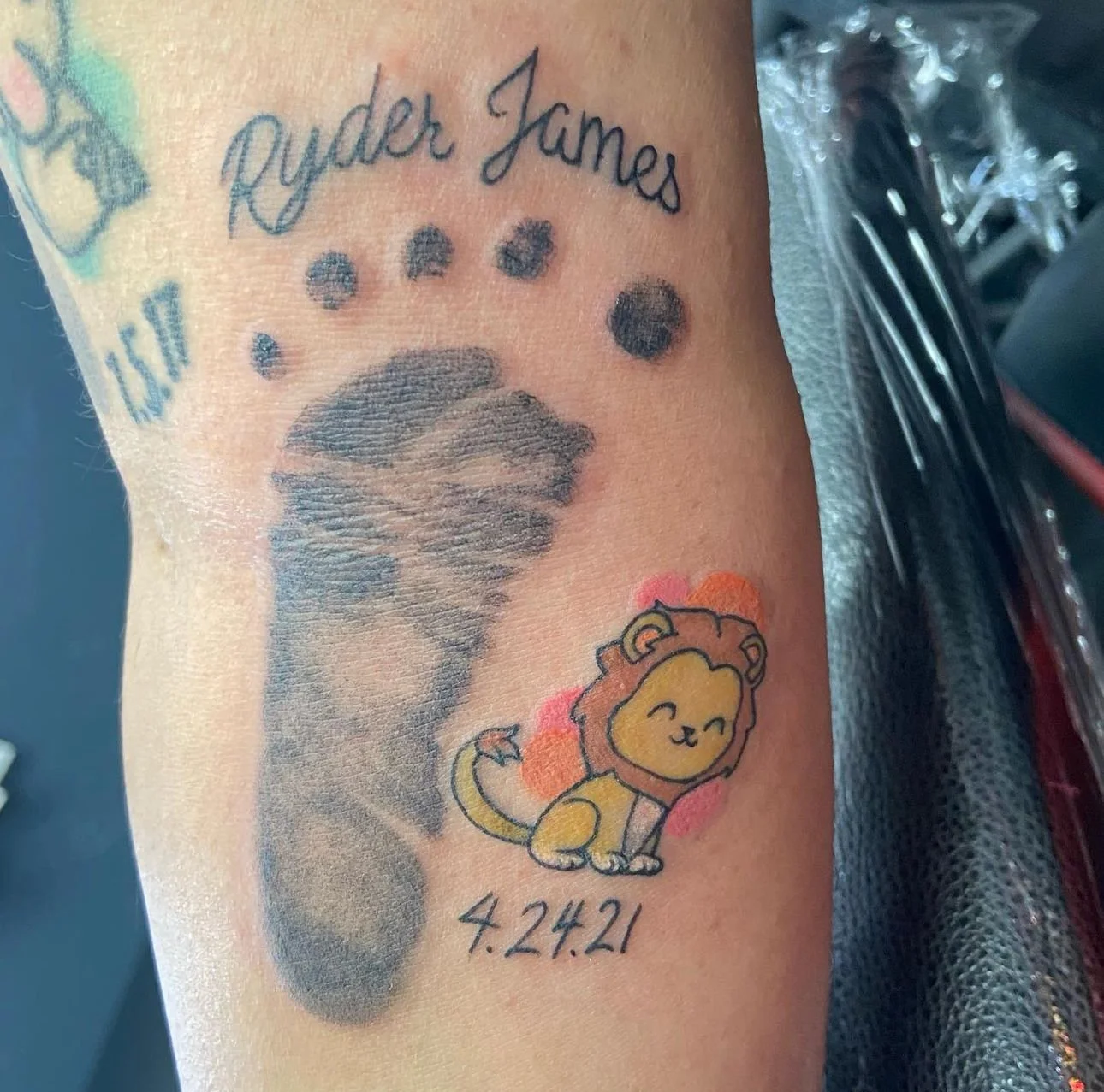 Tattoo of the name 'Ryder James' with six black dots, a whale, a cartoon lion, and the date '4.24.21' on a person's skin.