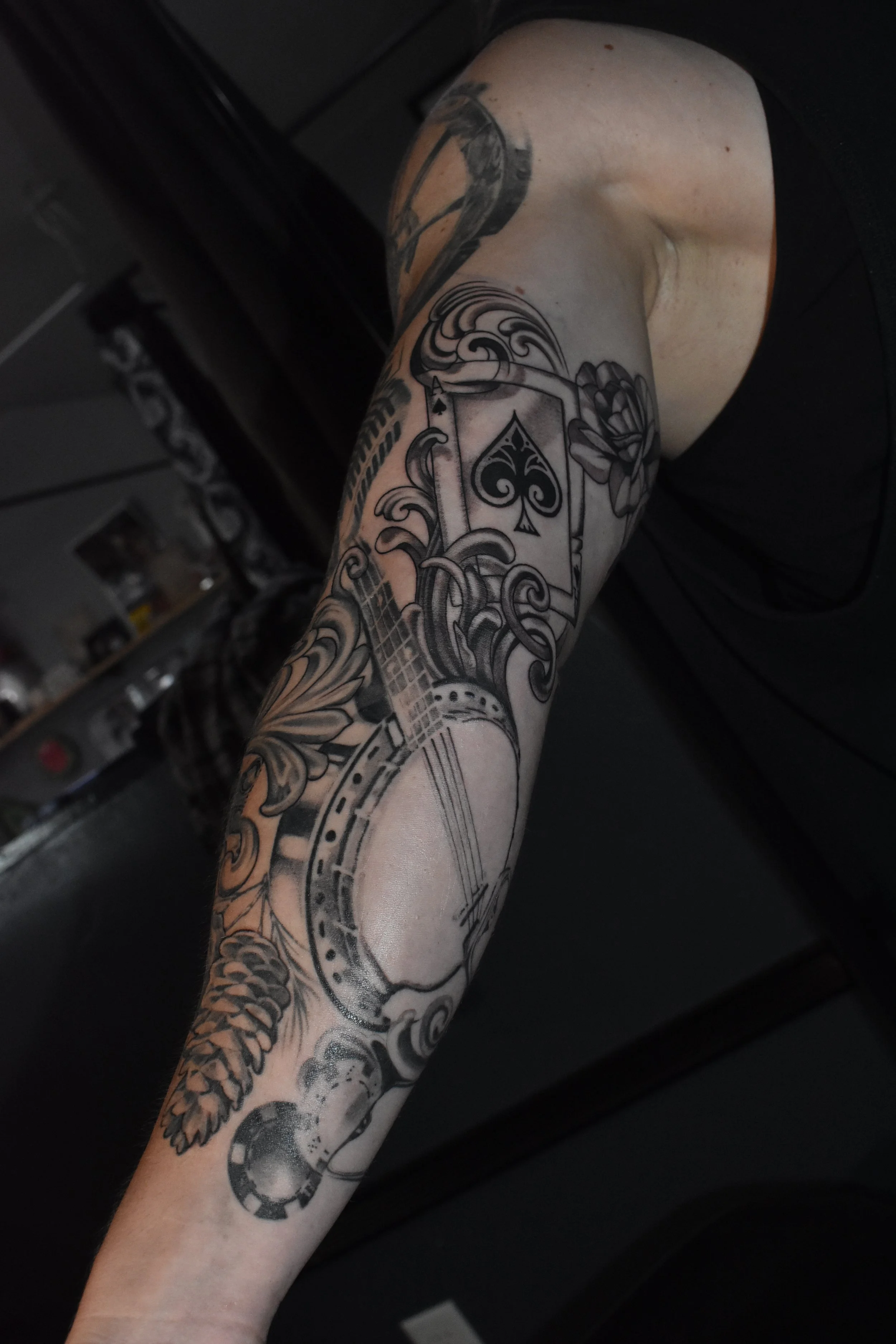 Black and gray tattoo sleeve featuring a guitar, playing card with spade symbol, clock, and floral elements.