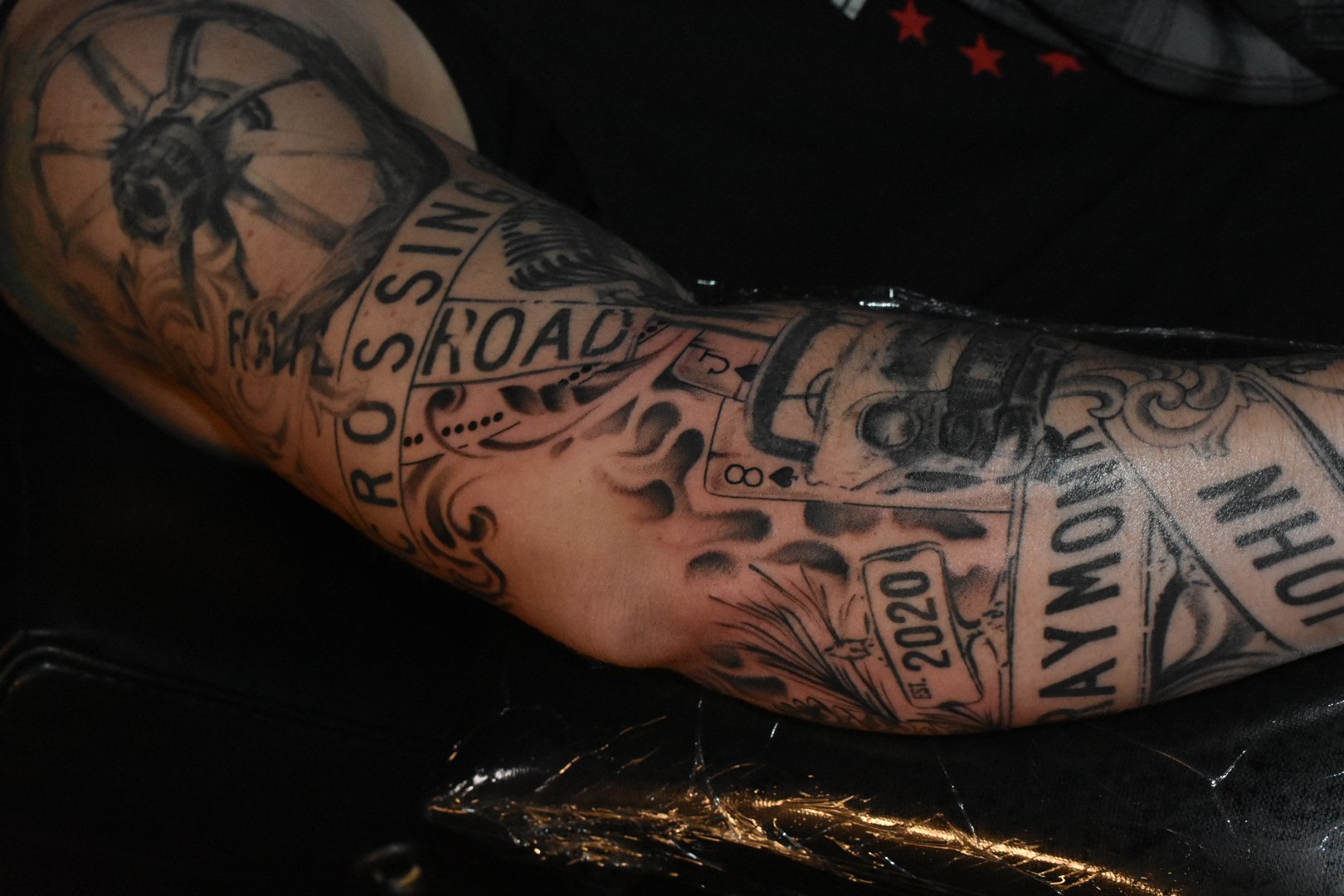 Tattooed arm featuring various black ink designs and text including a bee, a road sign, playing cards, and the year 2020.