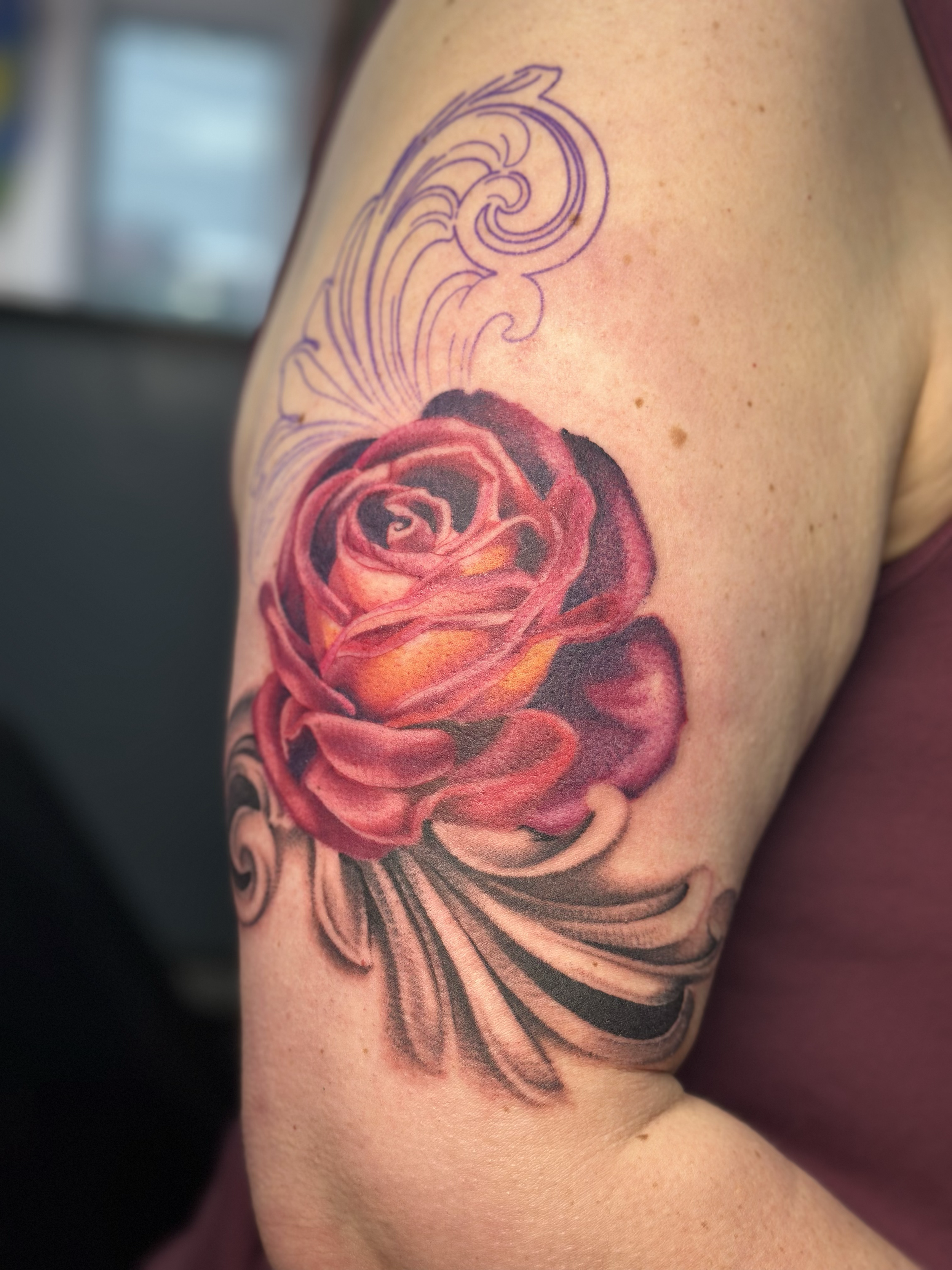 A close-up of a person's upper arm with a colorful tattoo of a rose in shades of red and orange, with black shading and detailed petals, and decorative swirling lines above it.