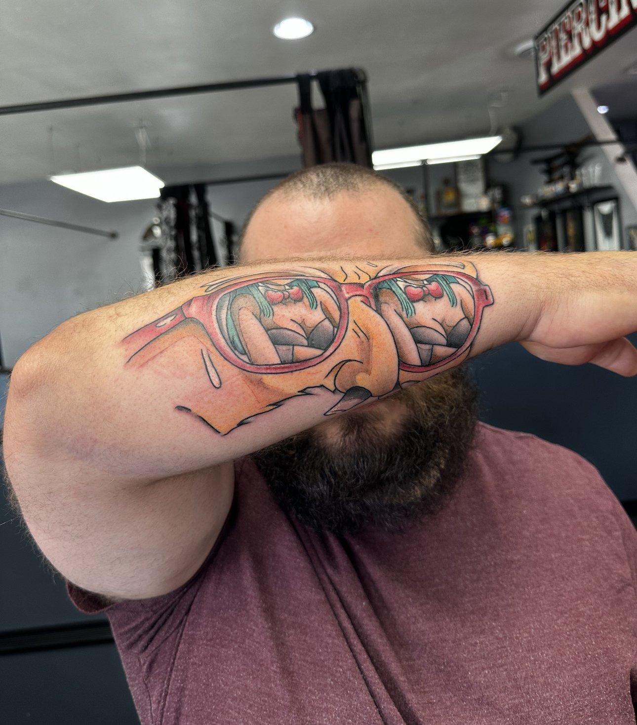 A man with a beard is showing a colorful tattoo of sunglasses on his forearm, which reflects an image of a cartoonish scene.