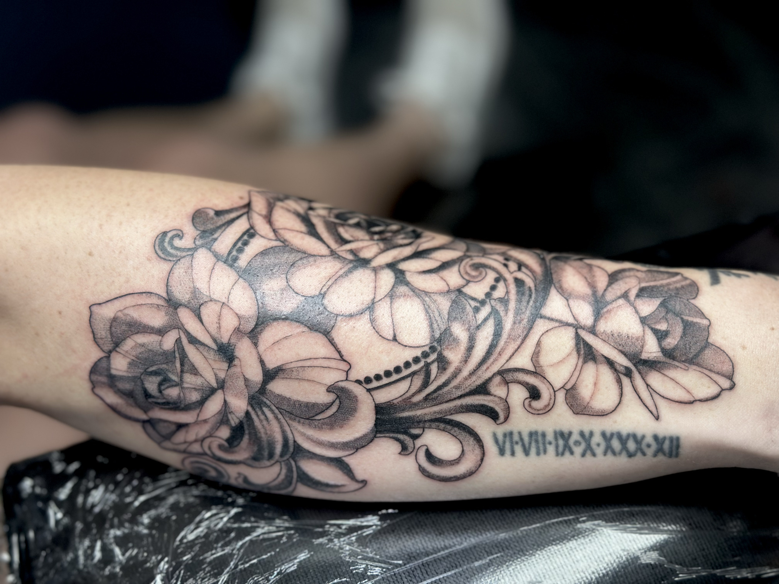 Close-up of a tattoo on a person's forearm, featuring floral and ornamental designs with Roman numeral text.