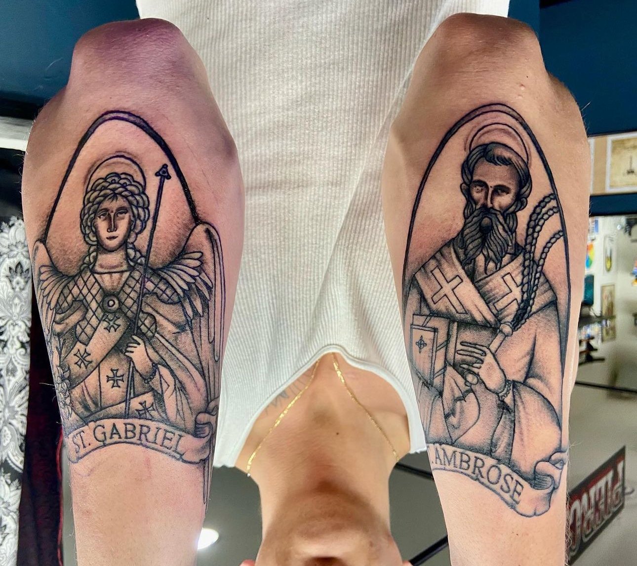 Tattoo of St. Gabriel and St. Ambrose on person's upper arms, featuring religious figures with banners bearing their names.