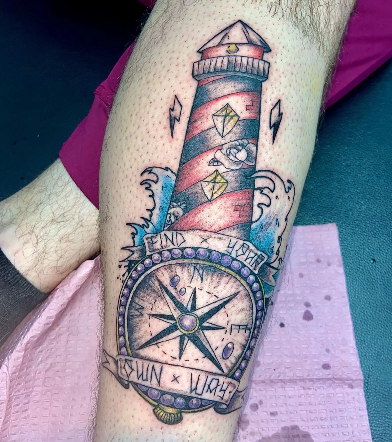 Tattoo of a lighthouse with red and gray stripes, clouds, two geometric shapes, lightning bolts, waves, a compass, and a banner with the words "Find Your Way."
