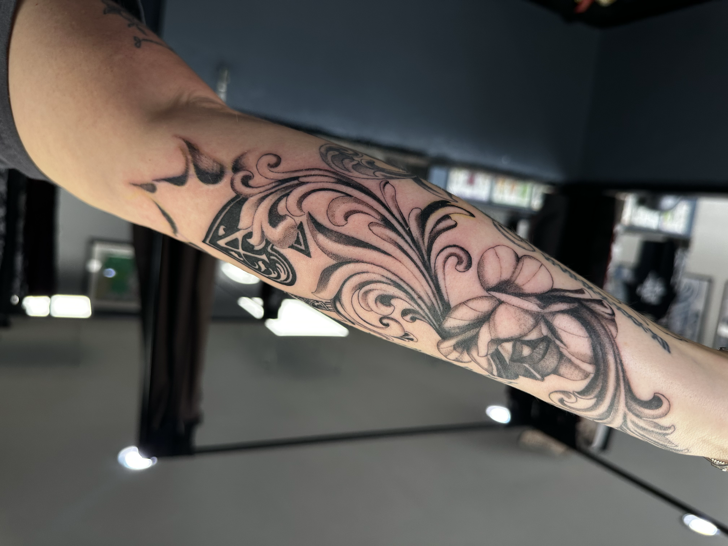 A person's arm with a detailed black and gray tattoo sleeve featuring floral and ornamental designs. The photo is taken indoors with a blurred background.