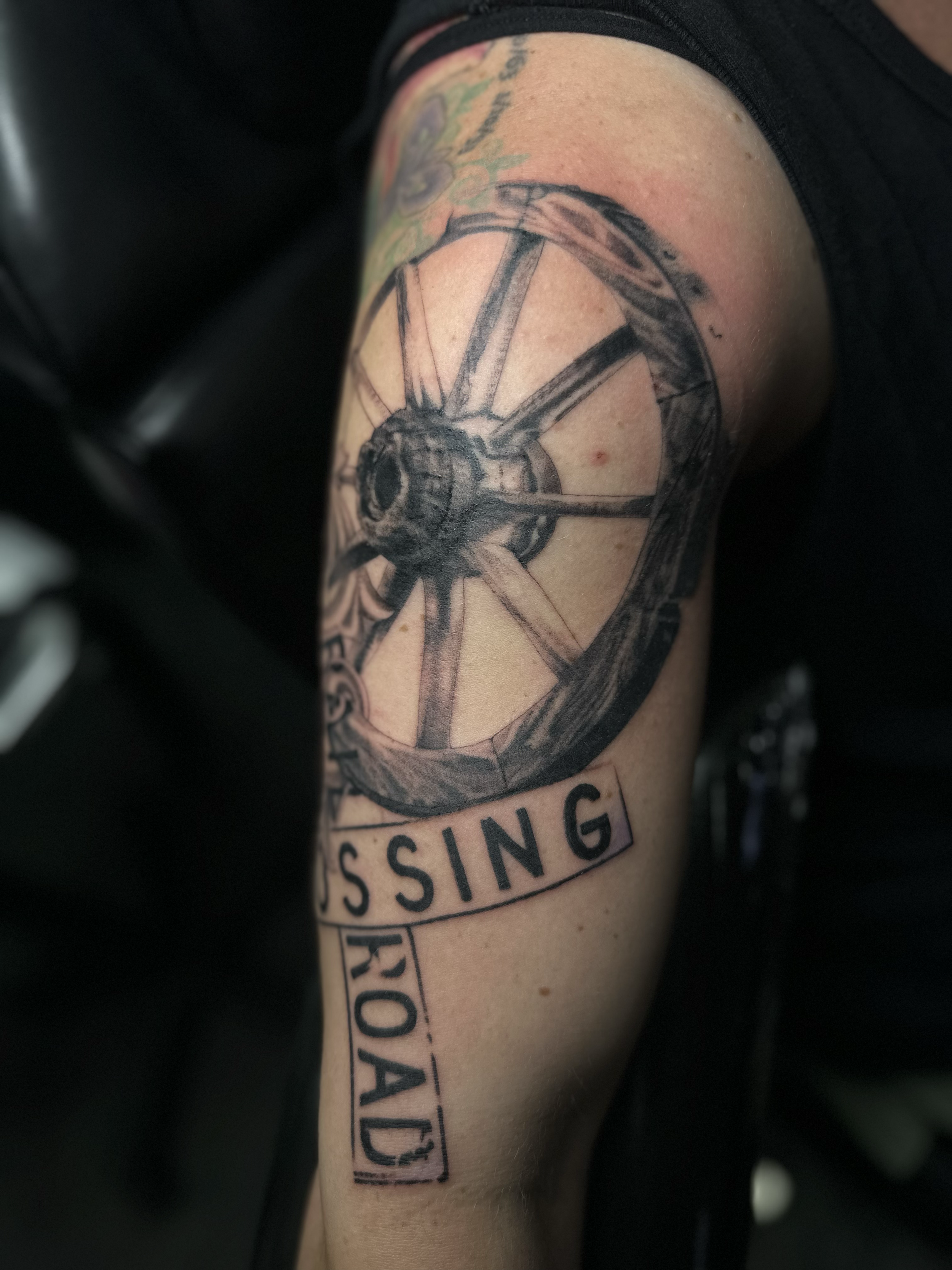 Tattoo of a bicycle wheel with spokes on a person's upper arm, with the words 'PASSING ROAD' in bold letters below.