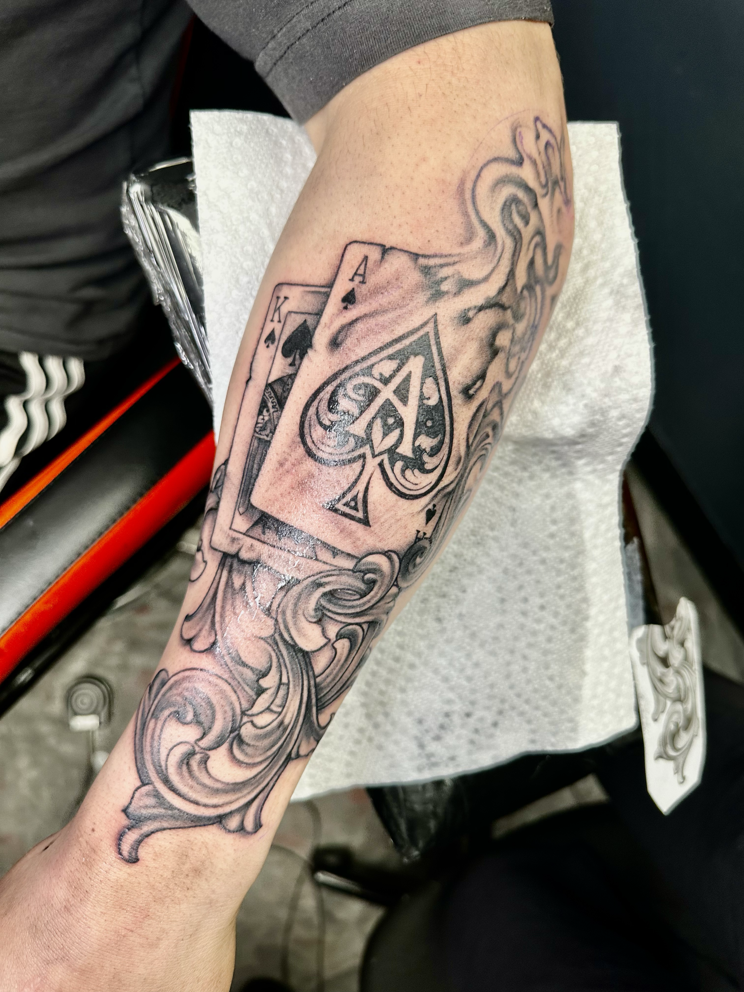 Tattoo of two playing cards, the King and Ace of Spades, with decorative swirling patterns on the forearm.