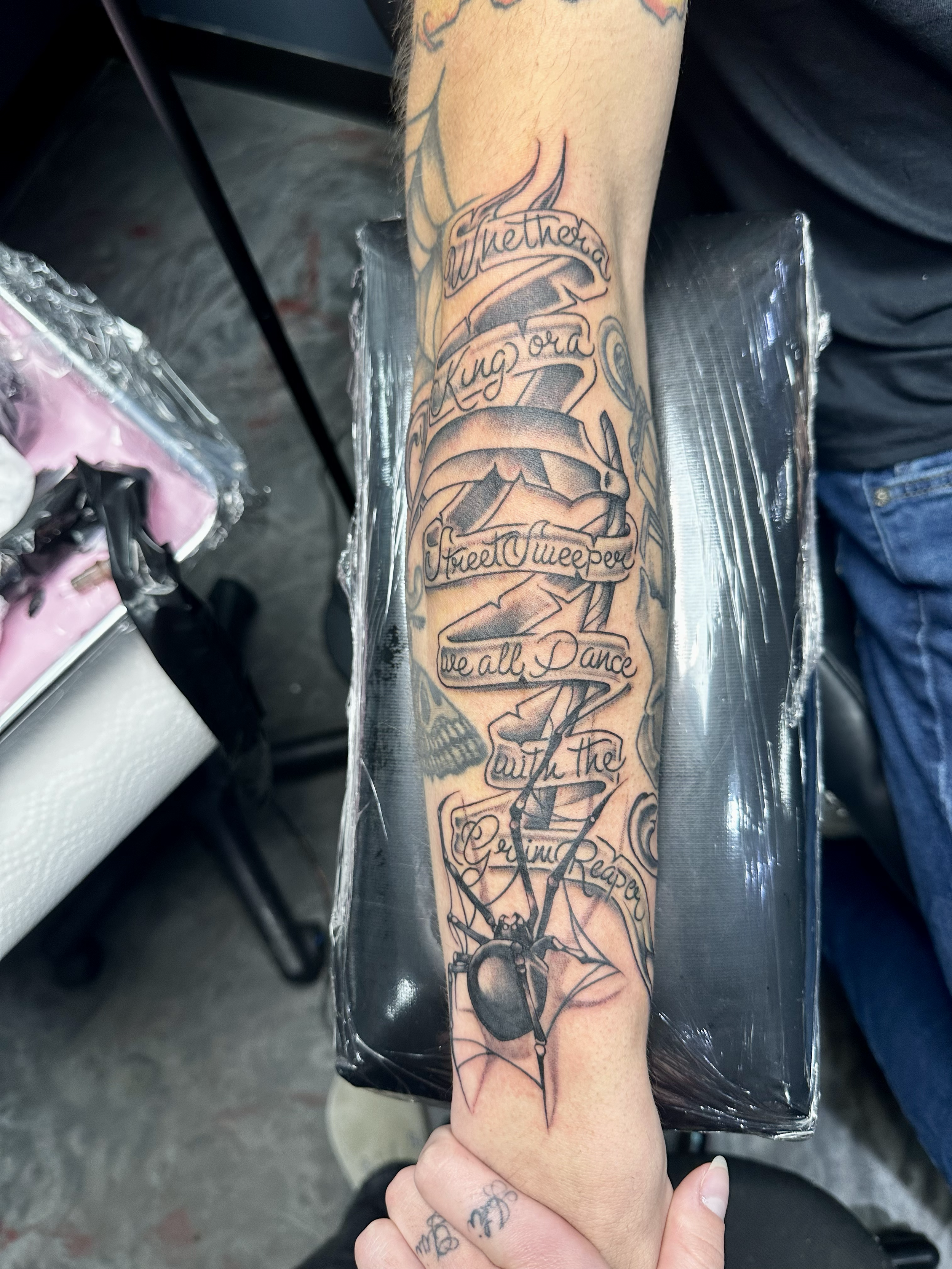 A tattoo on a person's forearm featuring a pendulum and banner with the words "Whether Kuga or Sweet Sneezer, we all Dance with the Rising Reaper."