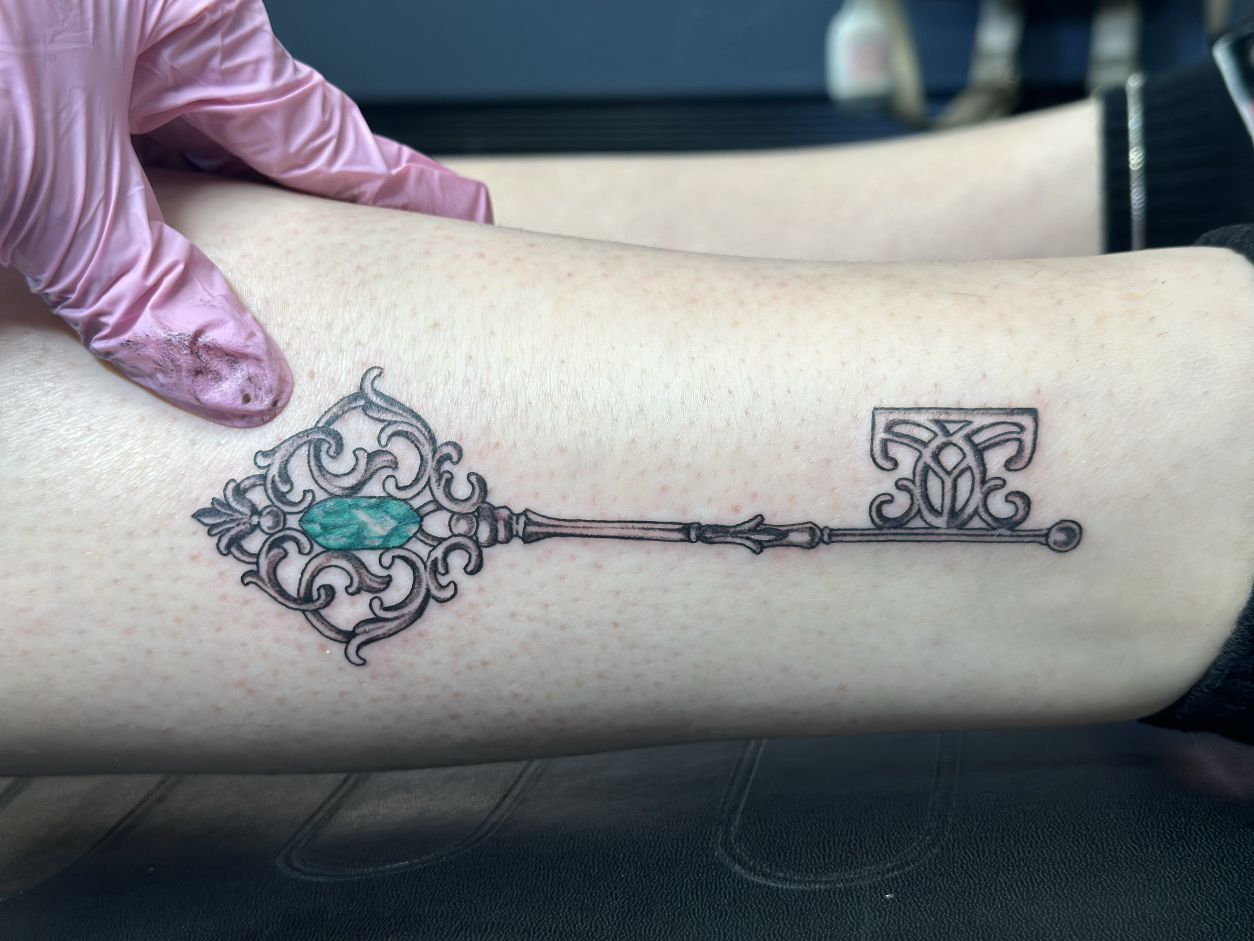 A tattoo of an ornate, vintage-style key with a turquoise jewel on a person's forearm.