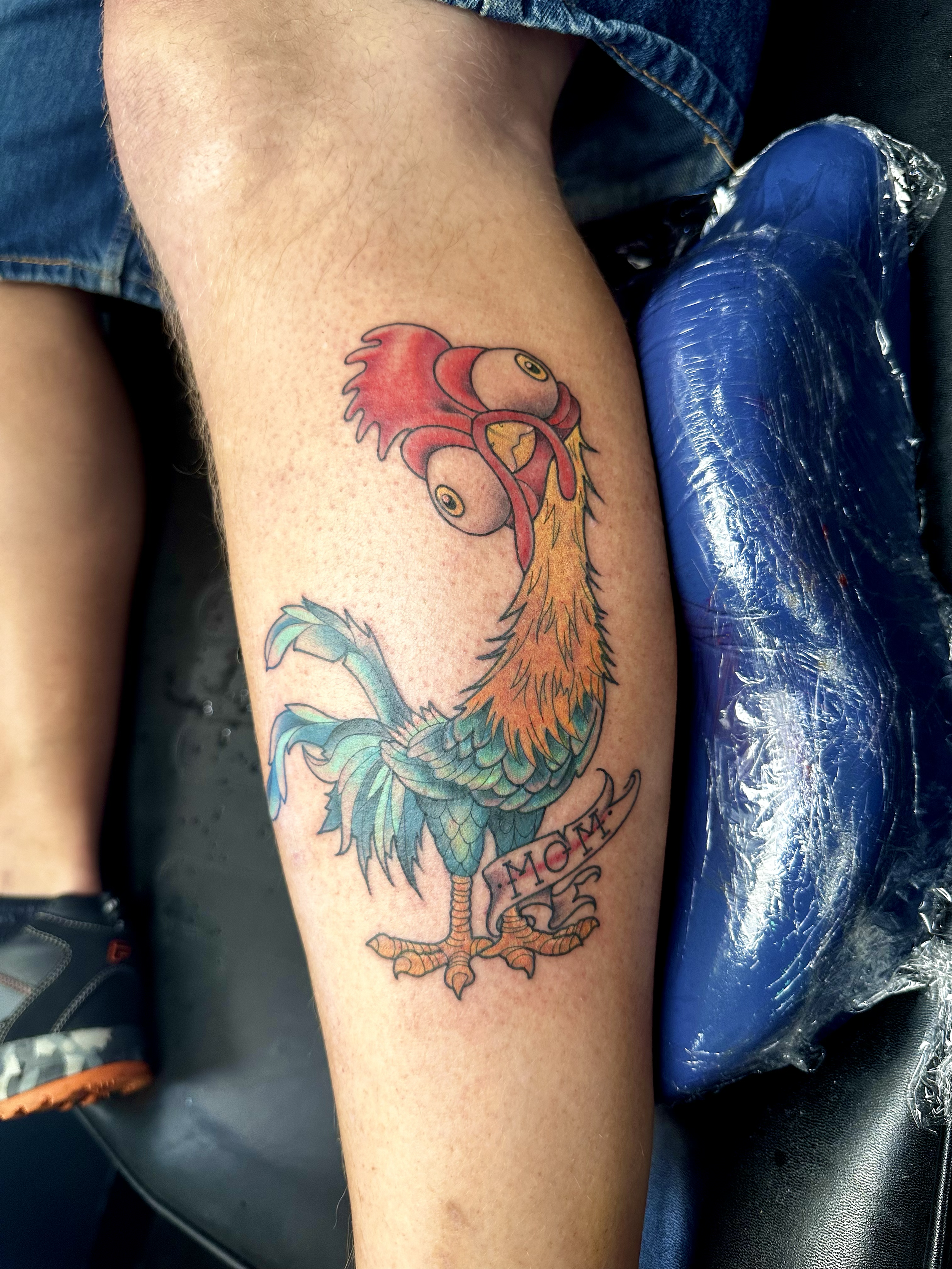 Colorful tattoo of a rooster with a banner that says 'Mom' on a person's thigh.