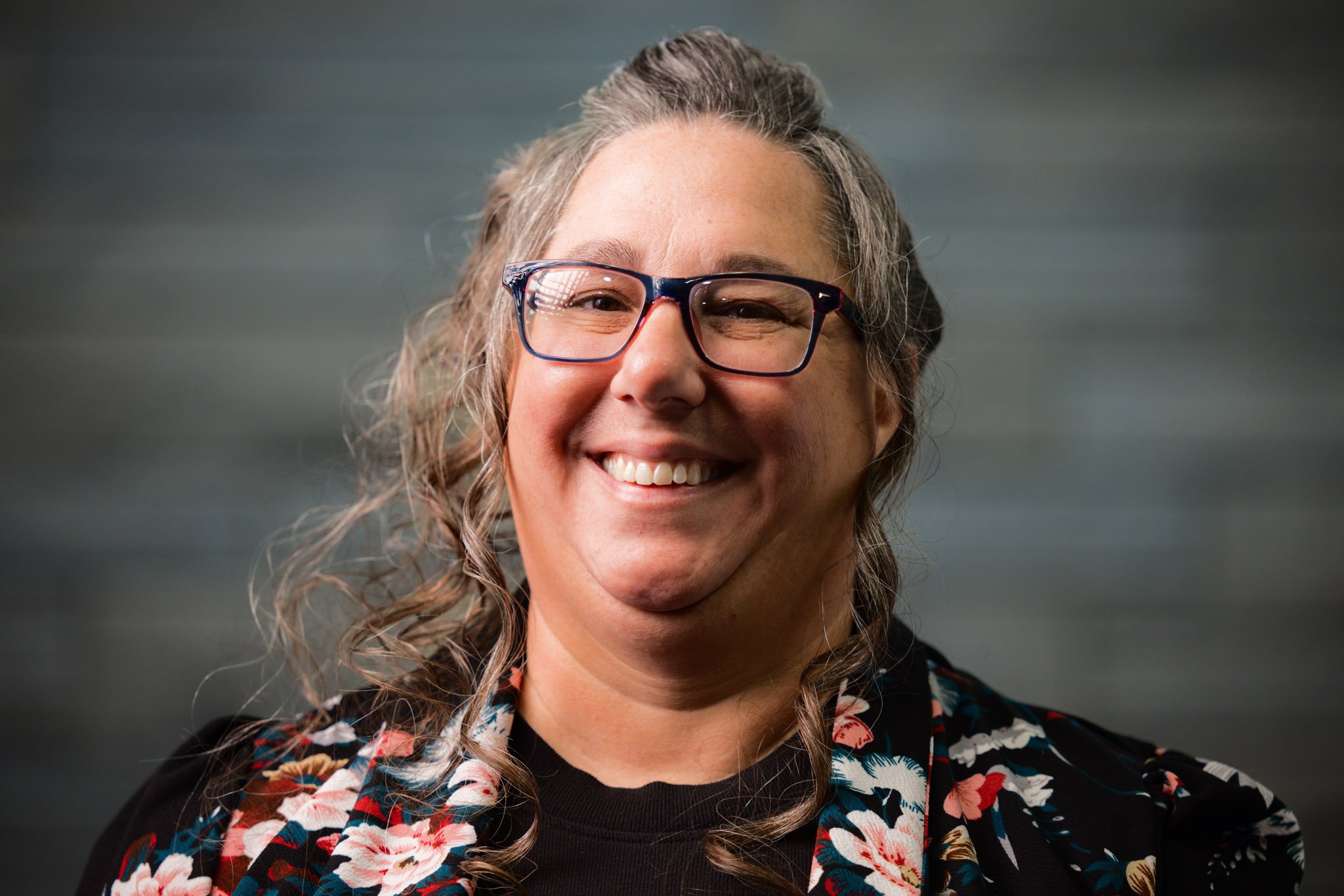 Close-up portrait of a smiling woman with glasses, curly hair, wearing a floral-patterned blazer, against a blurred dark background.