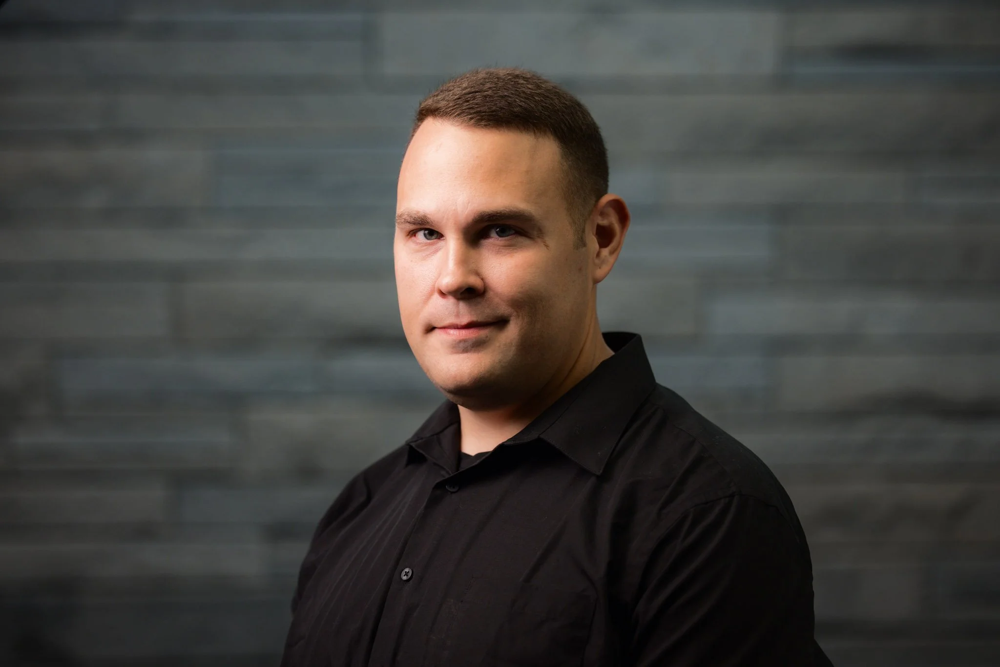 A man with short brown hair and light skin wearing a black button-up shirt against a dark gray wooden background.