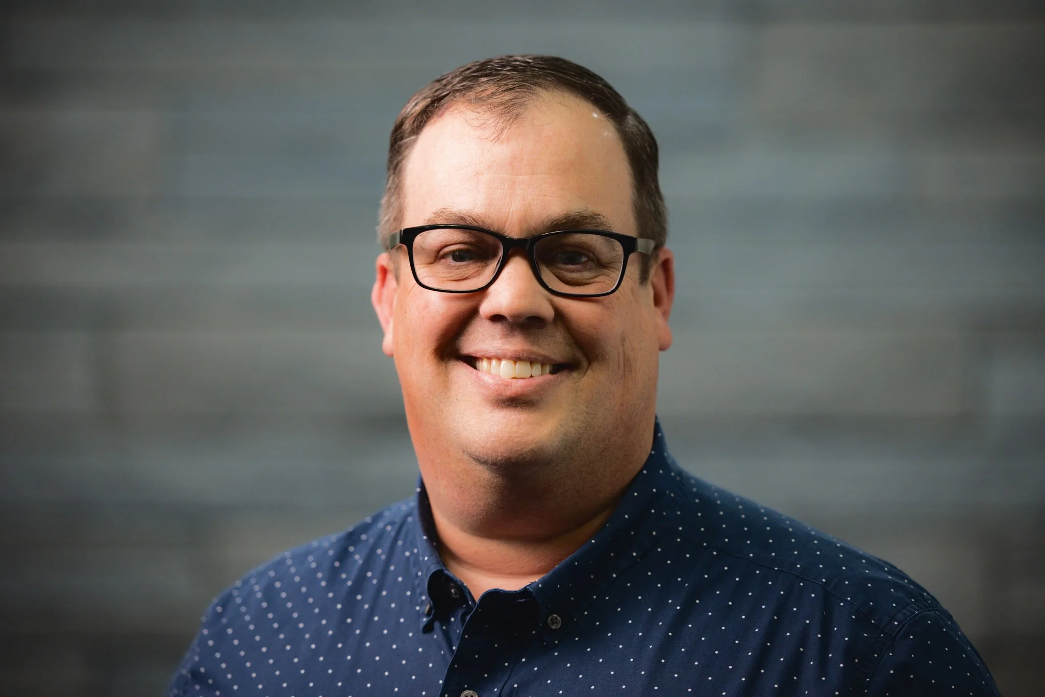 A smiling man with glasses, wearing a navy blue shirt with white polka dots, stands against a gray background.