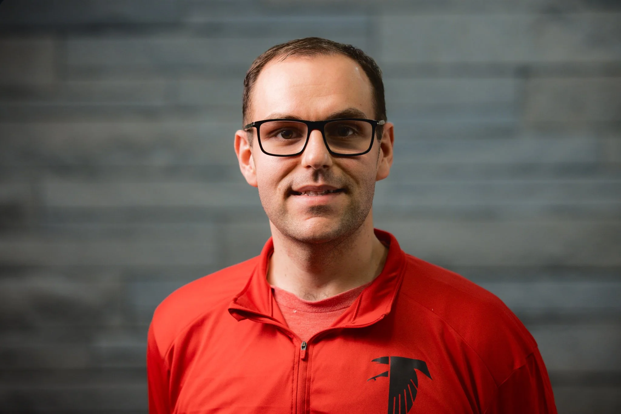 A man with short brown hair, wearing black-rimmed glasses, and a red jacket with a black logo on the chest, stands in front of a grey, textured background.