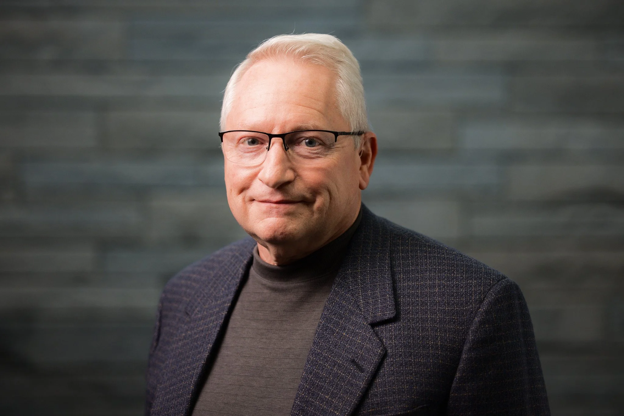 A mature man with glasses and short, gray hair, dressed in a dark blazer and turtleneck, smiling gently, standing in front of a textured gray background.