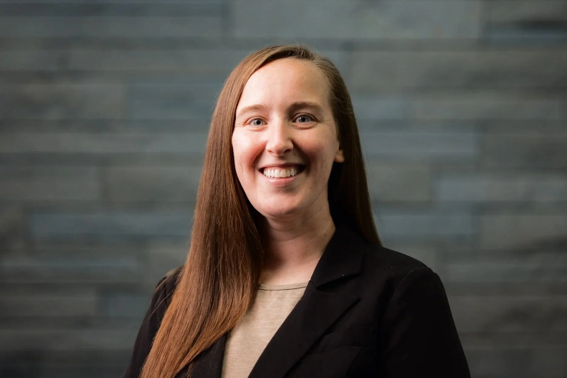 A woman with long red hair smiling, wearing a black blazer and beige top, standing against a dark textured background.