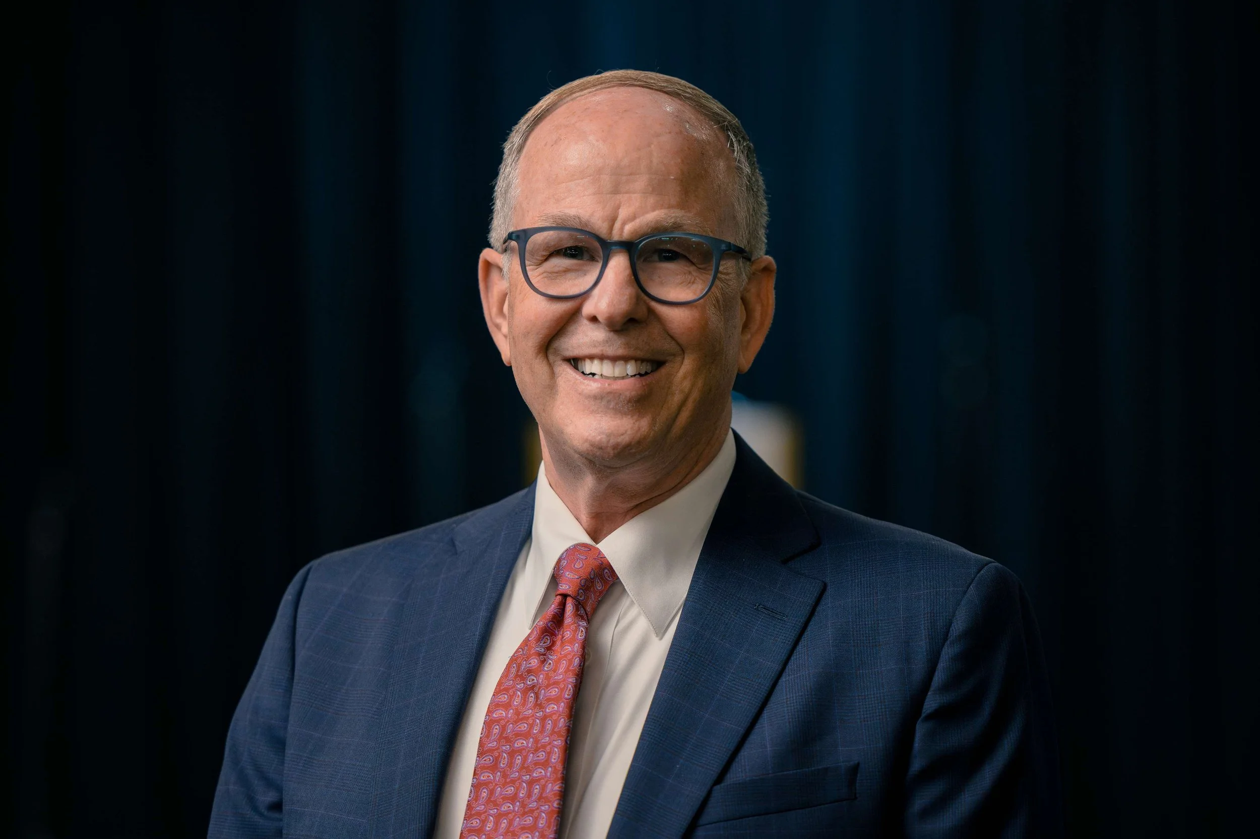 A smiling middle-aged man with glasses, wearing a navy blue suit, white dress shirt, and a patterned pink tie, standing against a dark background.